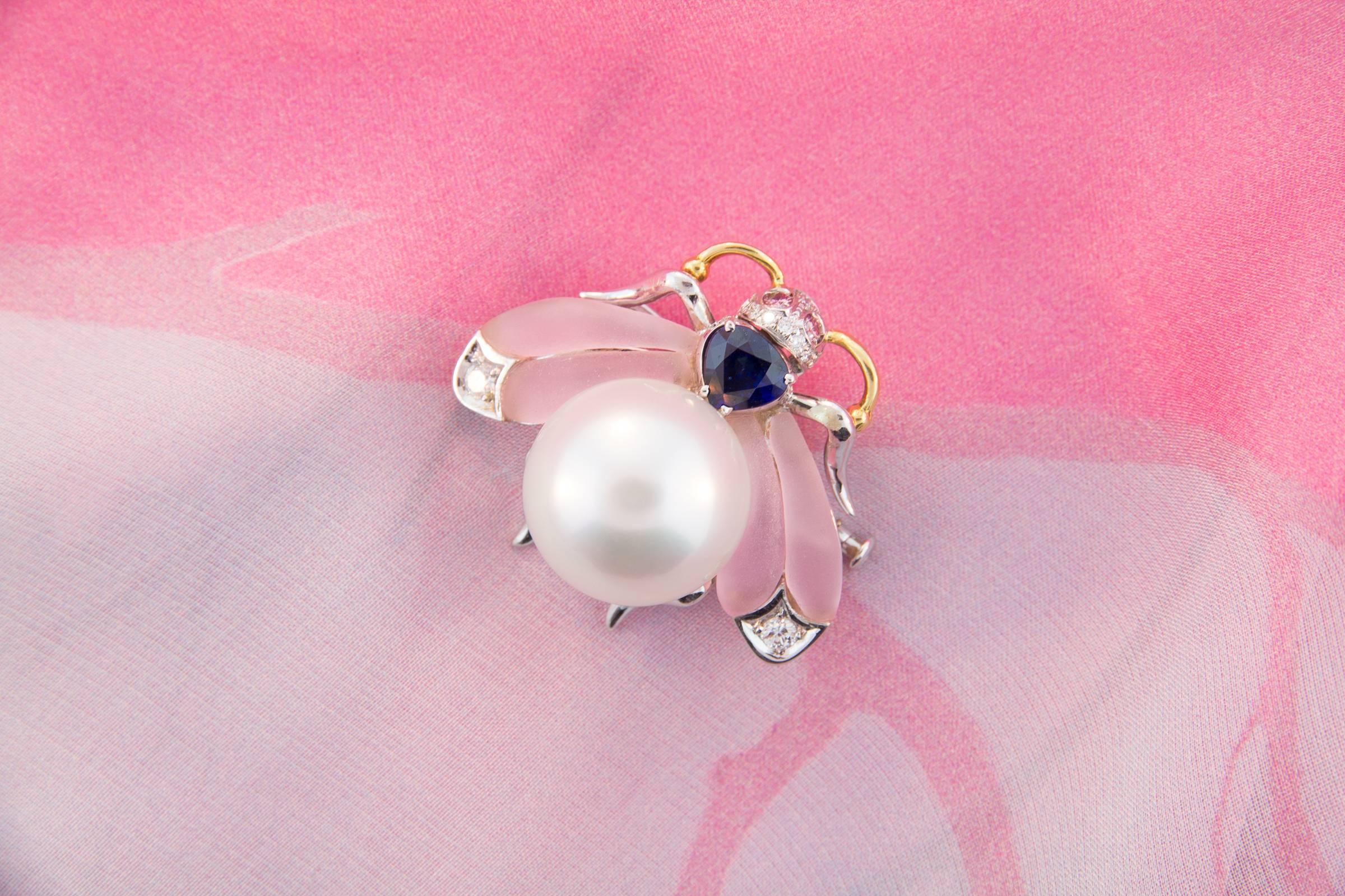This bee brooch features a South Sea pearl of 18mm diameter. The wings are set with tinted custom cut frosted crystal. The design is complete with a faceted drop shape blue sapphire (1.57 carats) and 0.30 carats of round diamonds. The eyes are