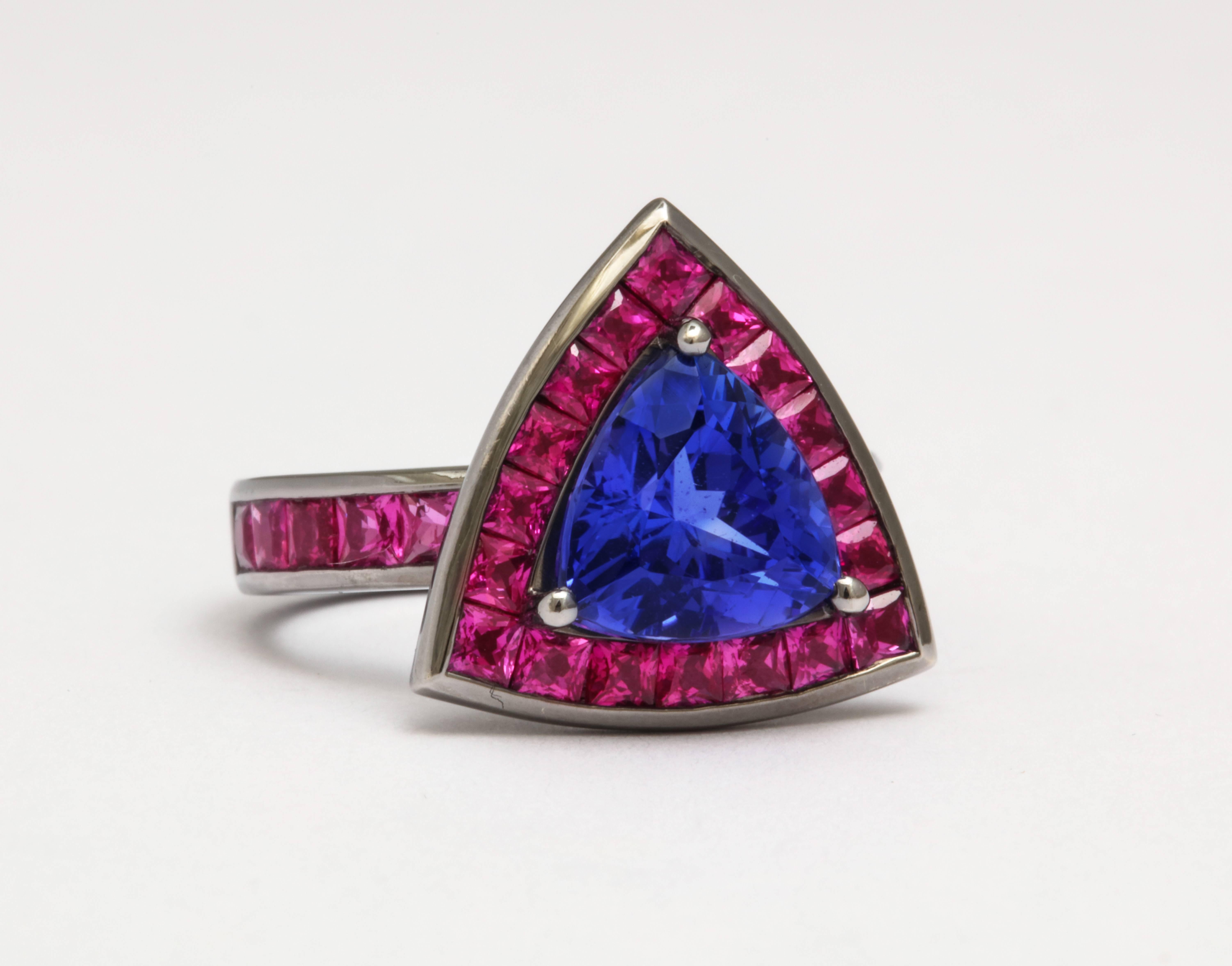 Dramatic and colorful 18 karat gold ring features a triangle cut tanzanite weighing 2.91 carats surrounded by 18 hot pink princess cut sapphires in a channel-set halo. Additional princess cut vivid pink sapphires are channel-set in the shank of this