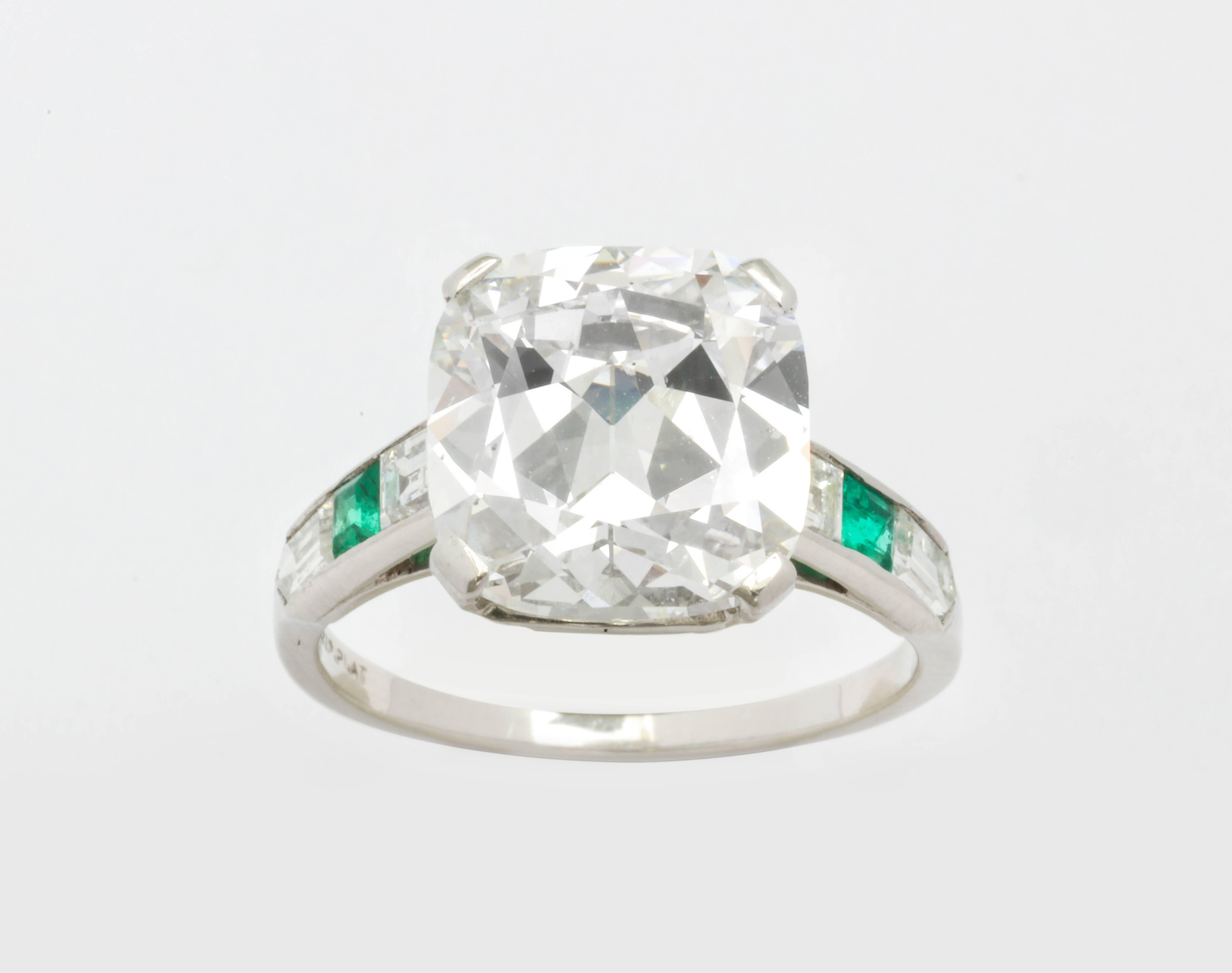 A diamond ring from the Art Deco Era featuring a 5.27 carat cushion cut diamond. Accompanied by a GIA certificate 2155593534 stating the diamond is an H color and SI1 clarity. The shank of the ring features two square emerald cut emeralds, two