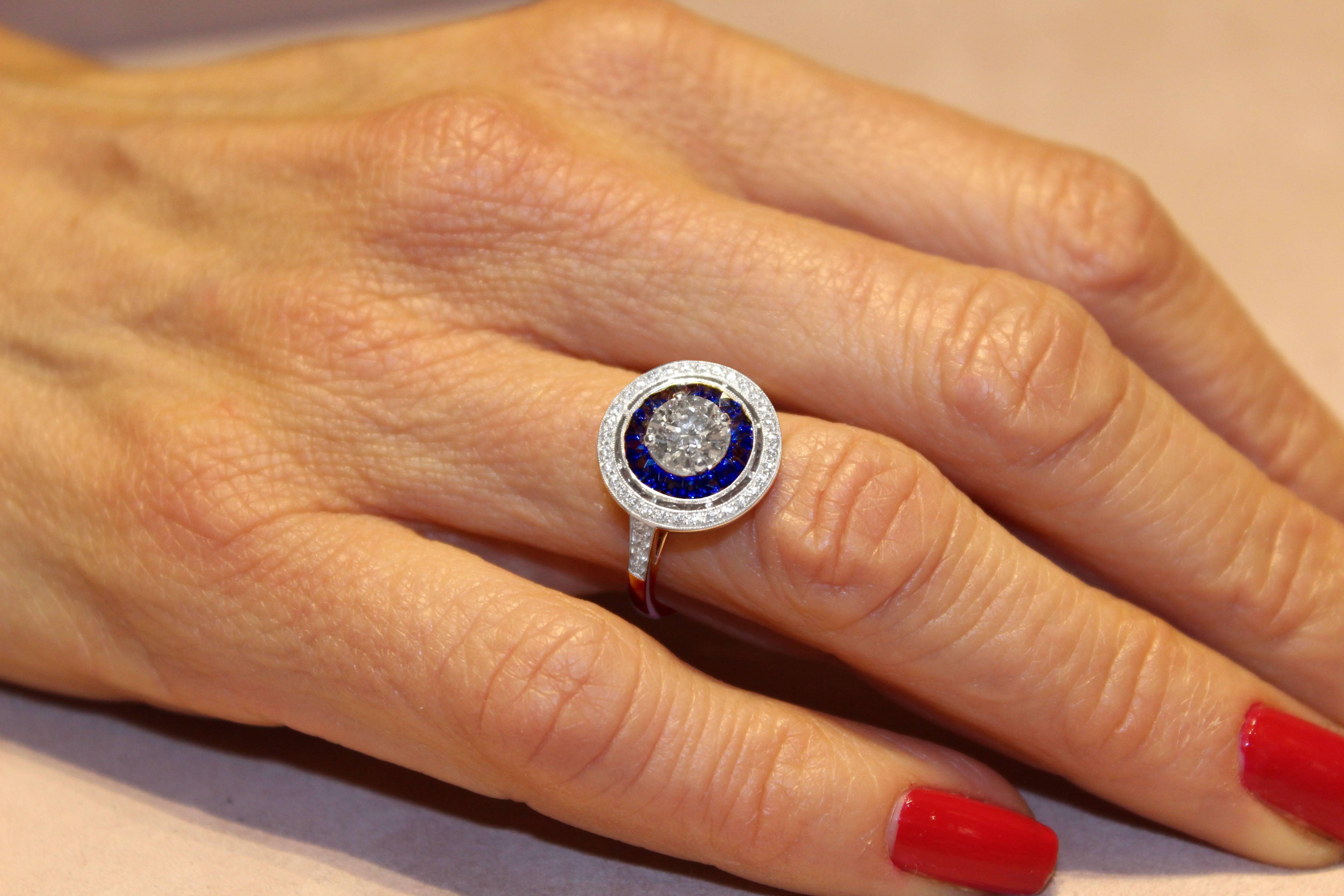 The 'Victorine' ring, designed in an Art Deco canted round shape is mounted in 18k white gold and showcases an exceptional 0.80 H VS1 brilliant-cut diamond, framed with a row of caliber cut sapphires weighing 0.50 carat, surrounded with a row of