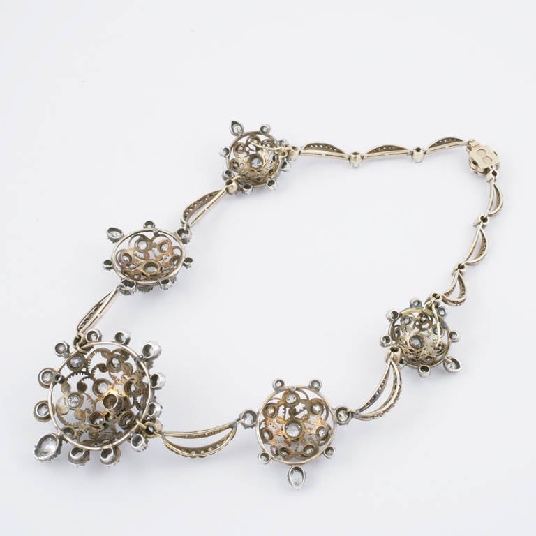 An Antique silver-top 15 karat gold necklace, composed of 5 openwork flower motif clusters.  The clusters are connected by diamond set swag links. Both clusters and links are set with old mine cut diamonds with an approximate total weight of 16.80