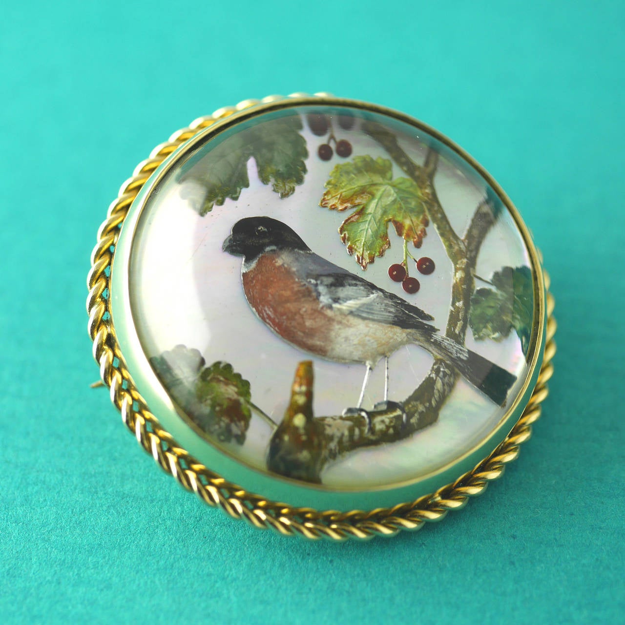 Essex crystal carved and painted showing a bird upon a cherry tree branch. Set on a mother of pearl background in 18kt gold. 

Essex Crystal or reverse intaglio are made from a cabochon (domed) crystal which has been carved from the back and then