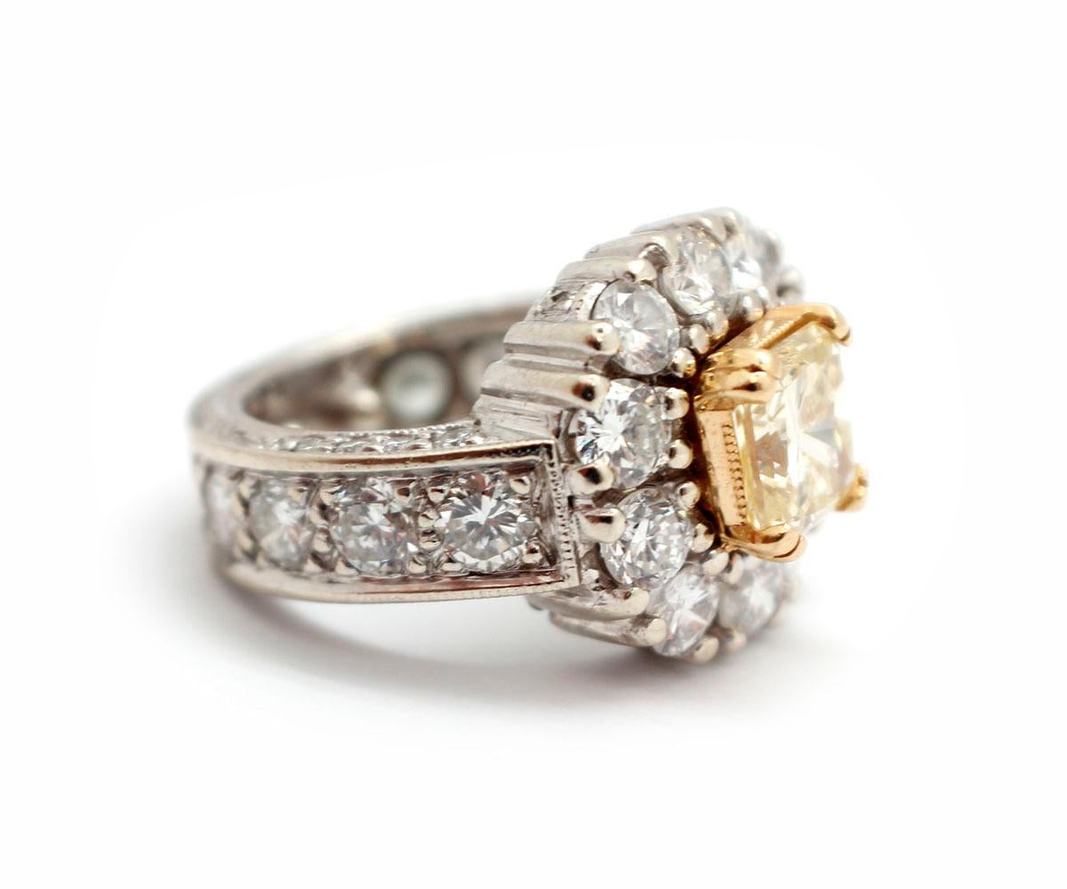 This stunning ring features a fancy yellow radiant-cut diamond in the center of a halo of round brilliant white diamonds. The center stone weighs 1.99 carats, and it is graded VVS2 in clarity. The surrounding diamonds have an additional weight of