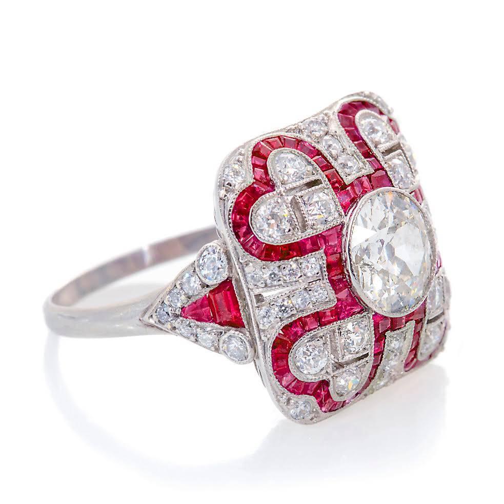 Featuring a bombe shaped, mill grained cluster with one, central bezel set, old cut diamond surrounded by a line of channel set, Carre cut rubies with the line extending to form heart shapes in each corner, within each heart are three, grain set,