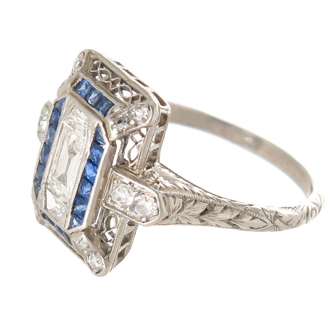 Circa 1930s Platinum, Diamond and Sapphire Ring, centrally set with a step cut diamond approximately 1/4 carat and flanked on either side by a step cut Trapezoid Diamond, further set with round diamonds and Swiss cut Sapphires. The Gallery and shank