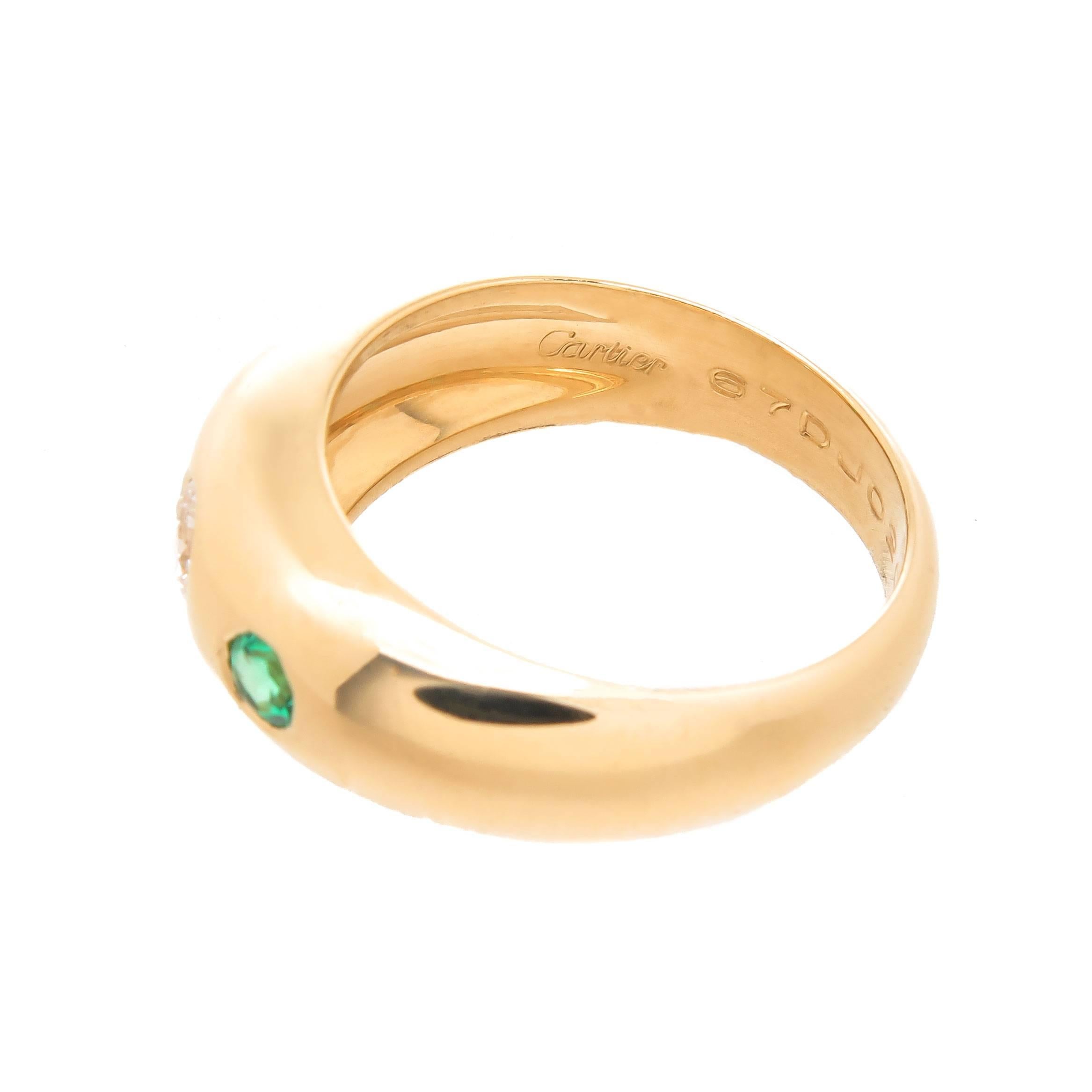 Circa 2000 Cartier 3 stone 18K yellow Gold Ring, centrally set with a Round Brilliant cut Diamond measuring 1/4 carat and grading as G in Color and VS in Clarity and having a very fine color Emerald on either side. The top of the ring measures 1/4