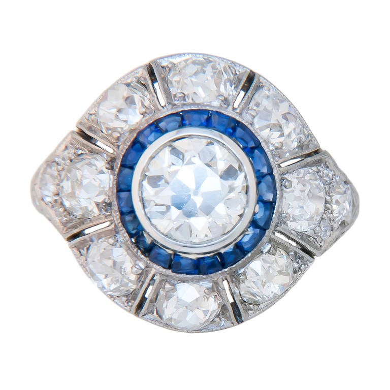 Circa 1930 Platinum and Diamond Engagement Ring, centrally set with a European Cut Diamond Weighing 1.13 Carat that is I in Color and SI in Clarity, surrounded by Swiss cut Sapphires. The ring is further set with old Mine Cut Diamonds that are very