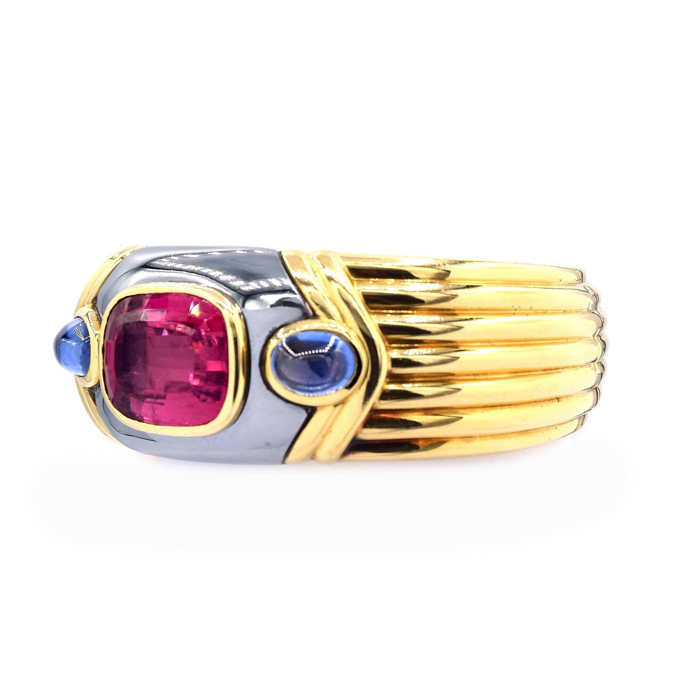 An Italian 18 karat yellow gold bangle bracelet with sapphire and rubelite by Bvlgari numbered BD 7499. The open back flexible bracelet has 2 cabochon sapphires and 1 rubelite. All three stones are bezel set in the bangle. 
Signed 