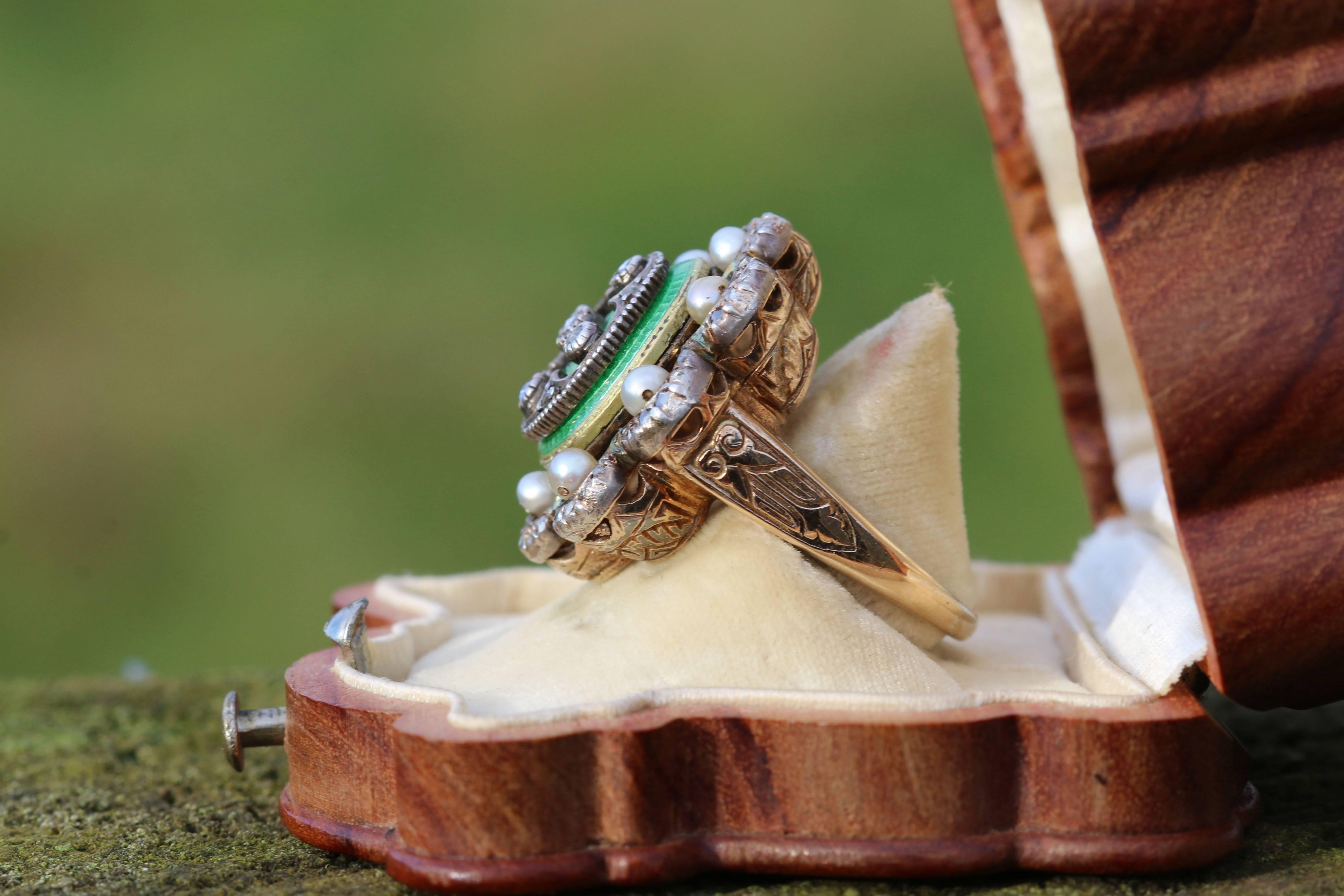 This is a beautiful and unusual green enamel ring with pearls surrounded by rose cut diamonds in a stylised flower design. The ring is gold with the diamonds set in silver. It is marked 