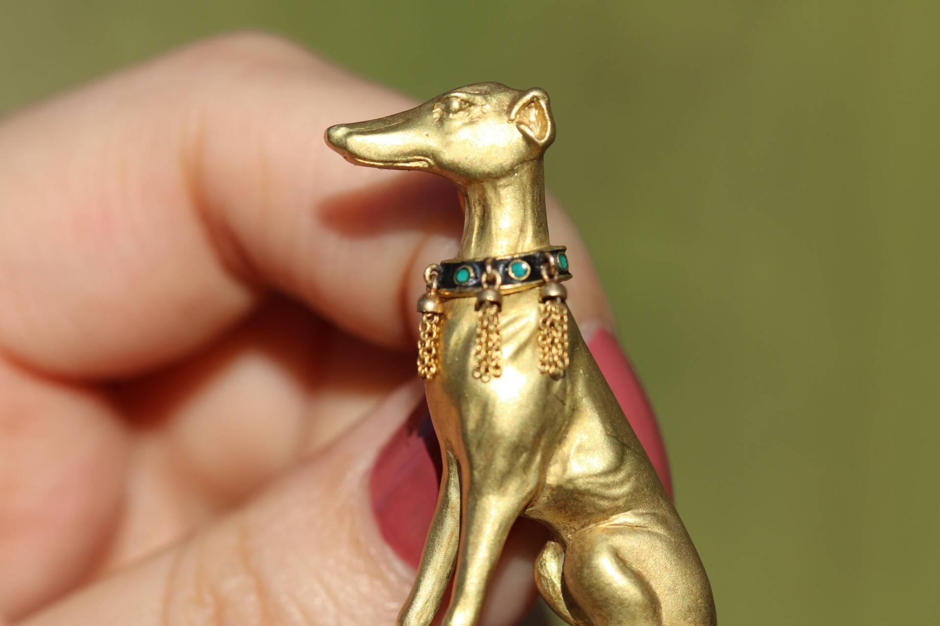 This brooch shows the sleek and elegant form of a greyhound sitting on a platform lined with diamonds over an Art Nouveau inspired flourish of flowing gold. Below the platform are suspended three pear-cut emeralds. The Greyhound is wearing a collar