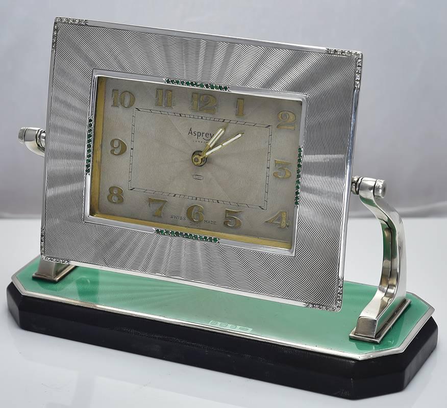 Elegant art deco sterling silver desk clock.  Made and signed by ASPREY.  The frame is beautiful, with a wide engine-turned silver border, set with emeralds around the face and diamonds on the outer corners.  Applied gold numerals.  The base is