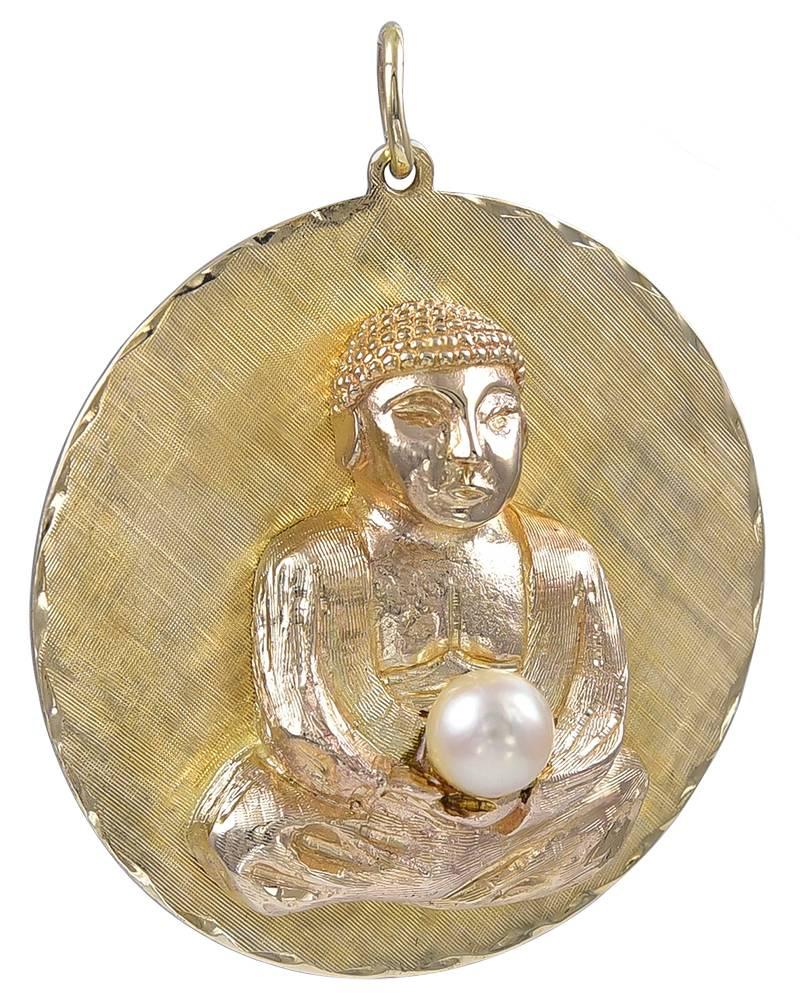 A great charm:  a huge pendant with a three-dimensional buddha.  The buddha is seated and is holding a pearl with both hands.  Set on an engine-turned background, with a beveled edge.  14K yellow gold. 1 5/8" in diameter.  A rare subject,