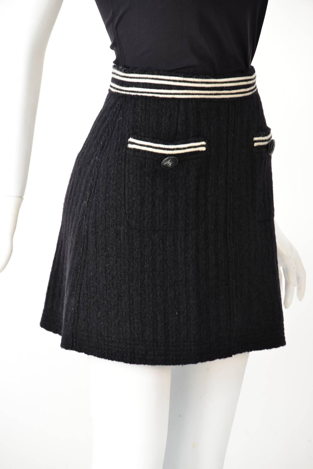 Minigonna moderna degli anni 2000 in lana nera di Chanel. La gonna è dotata di una fascia in vita con finiture bianche e nere a contrasto e di due tasche anteriori da lavoro. Un bottone con il logo di Chanel è presente in ogni tasca. La gonna si