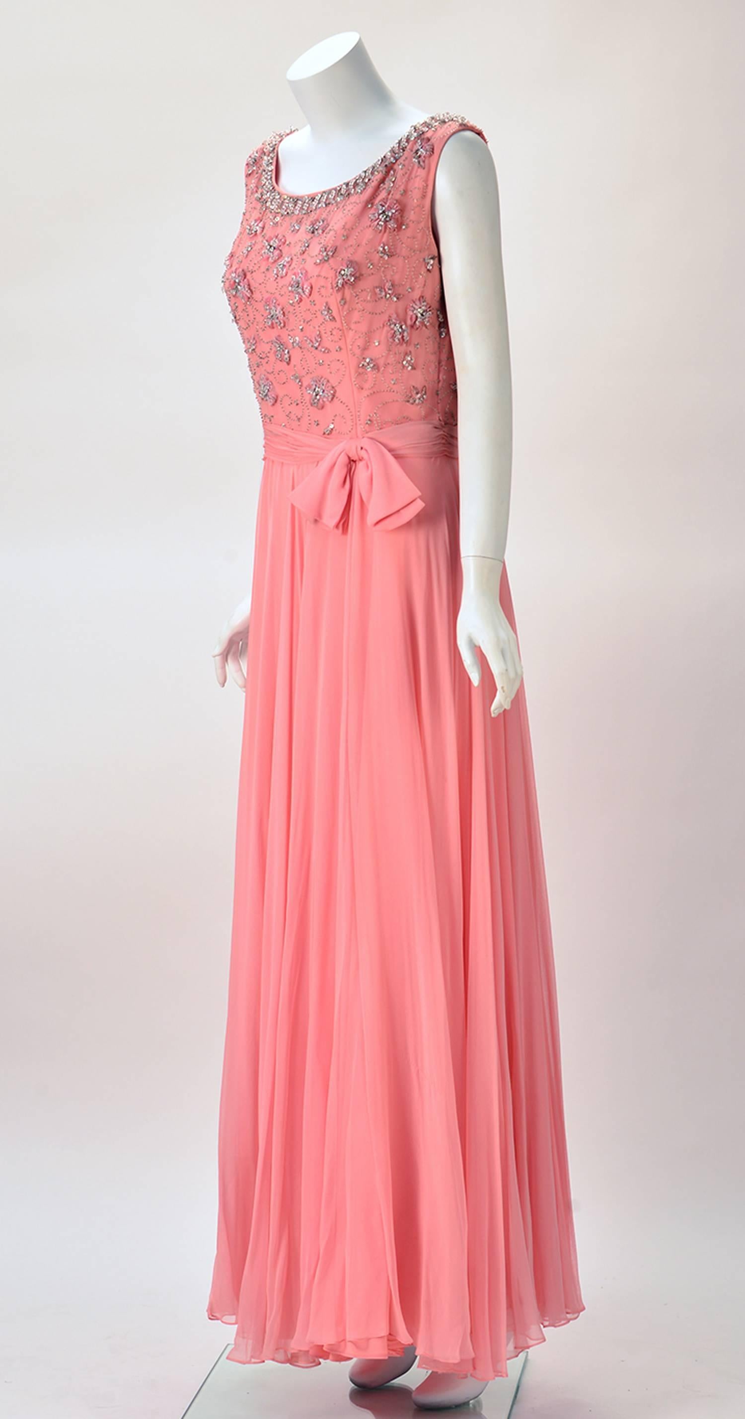 1960s bubblegum pink sleeveless evening dress by Cameo Evening Fashions. The dress features a beaded floral scene bodice with embroidery that boasts silver metallic and pink velvet threading. A gathered waistline with bow offsets the flowy layered