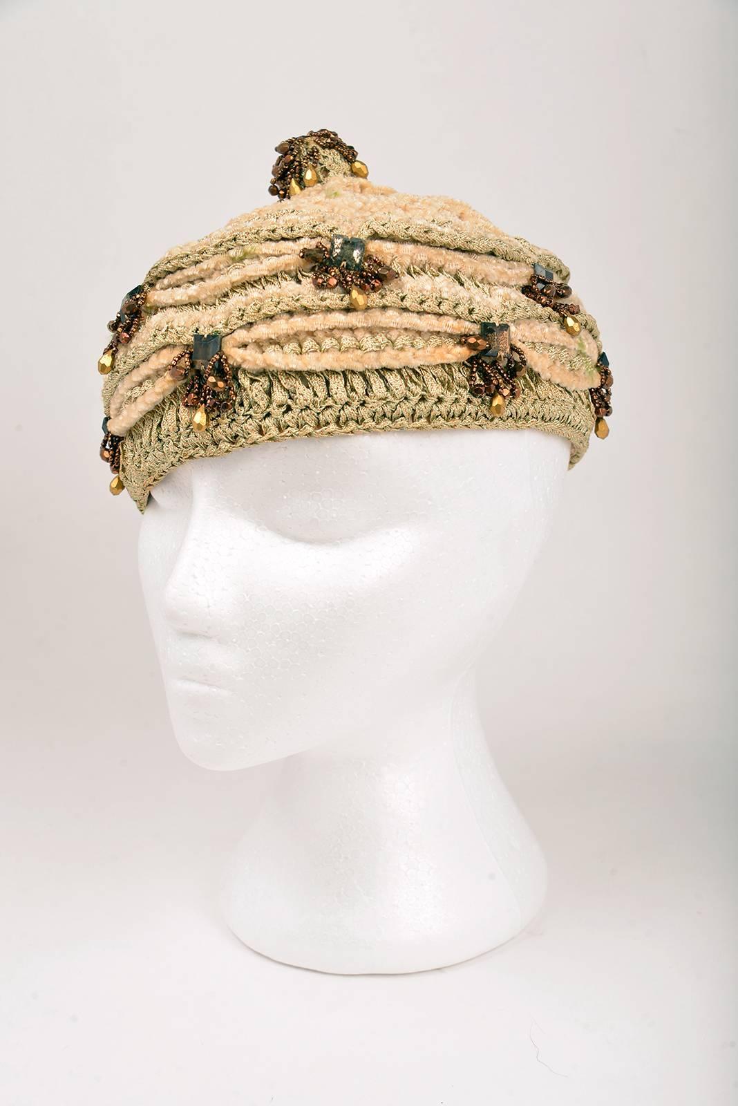 A delightful vintage hat by Gladys and Belle! This structured beanie is composed primarily of ecru wool yarn, peach ribbons arranged in a bunting-like manner, and several beaded appliques. The beaded appliques feature square-cut jewels, surrounded