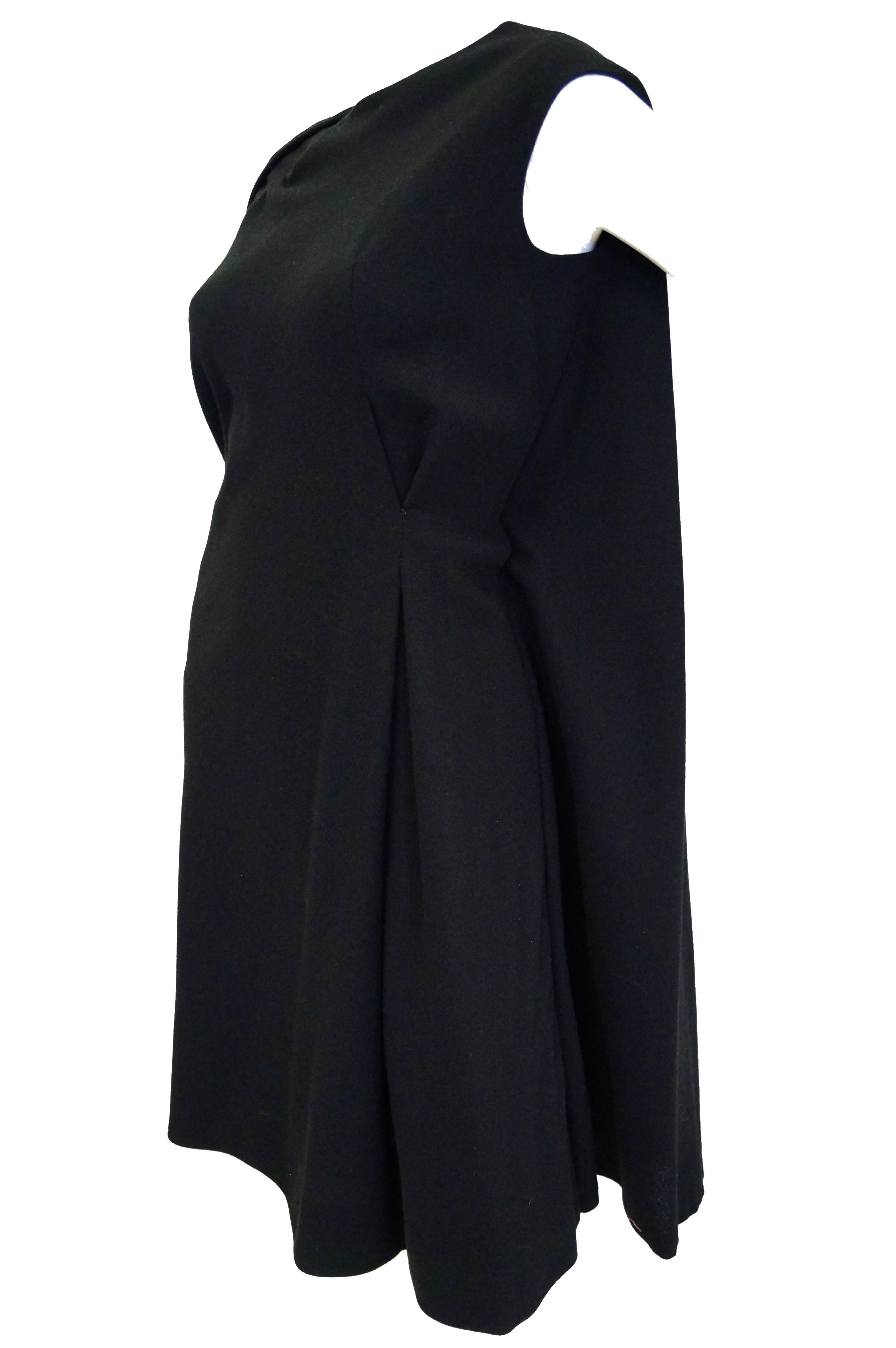 Fantastic black wool cape dress by John Moore! This dress sits just above the knee, has a jewel neckline, and short sleeves. The loose shift silhouette of the dress is interrupted by a fine and precise pleat on the hip. The dress has a fabulous cape