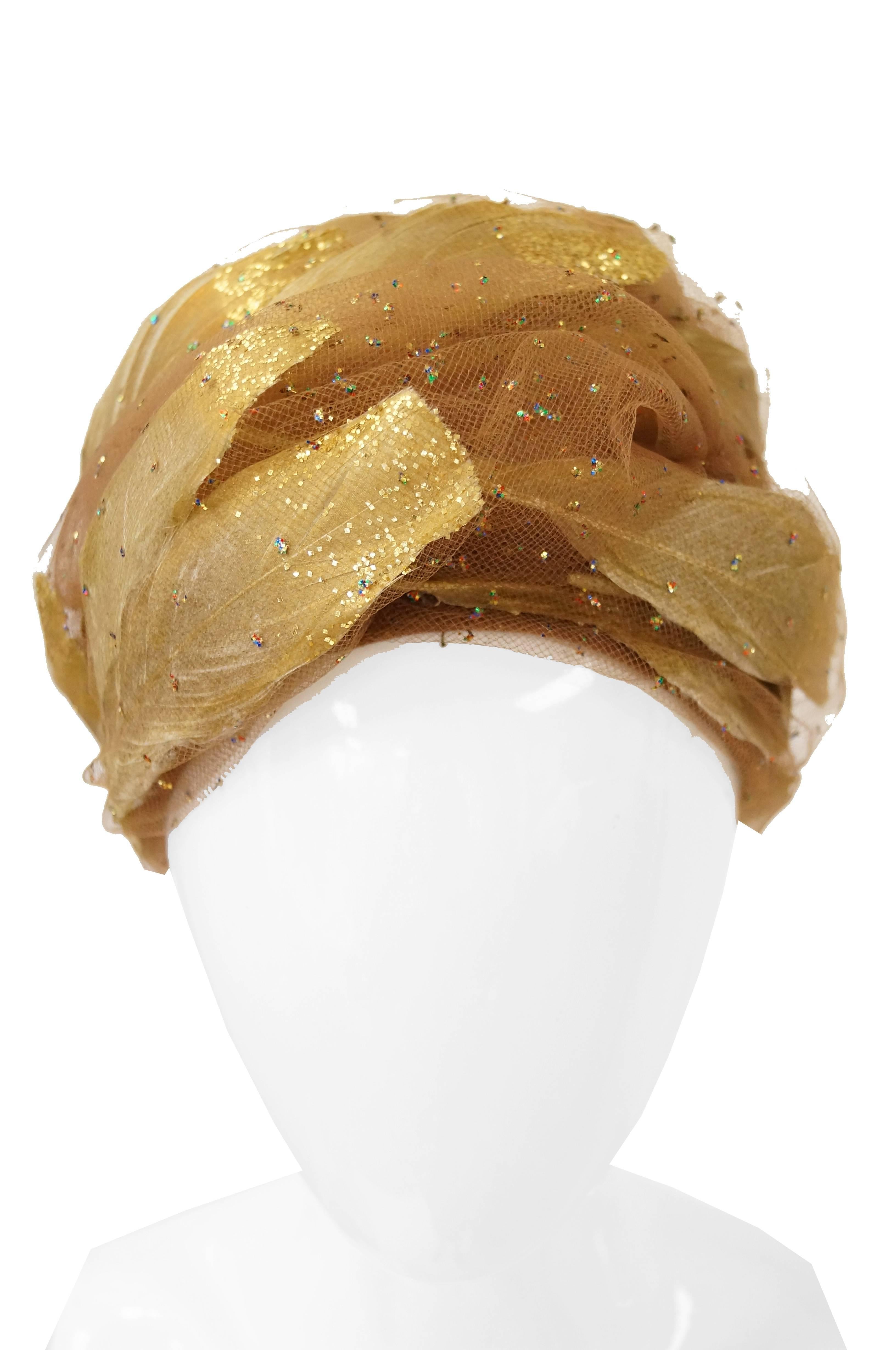 From a distance the toque hat appears to look like a golden rose on the brink of full bloom. When seen up close one can distinguish the feathers from the tulle and glitter, making the wearer of the hat seem as though their head is being cradled by