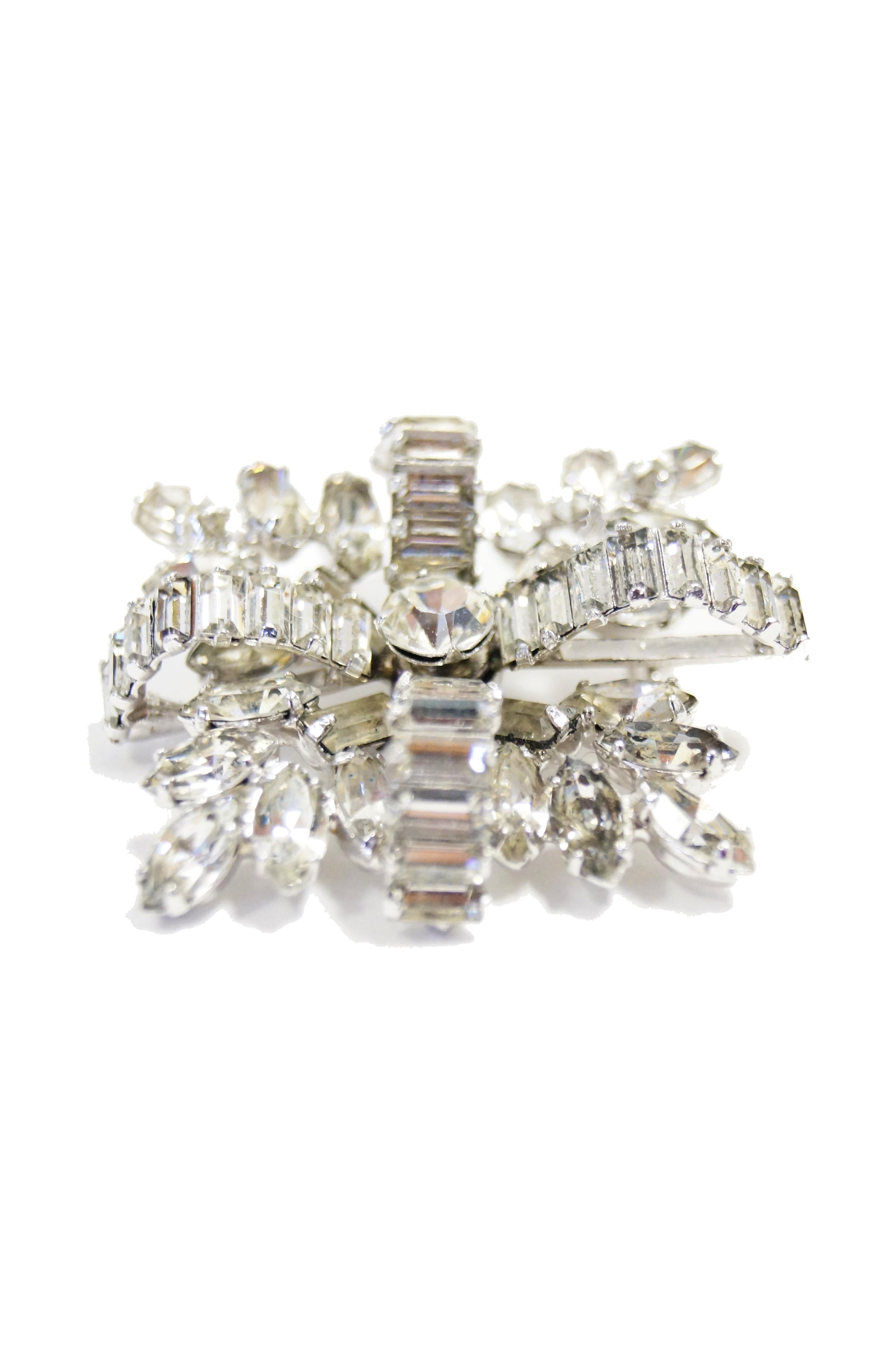 Gorgeous clear and bright rhinestone brooch by Hattie Carnegie. This brooch has a round, multifaceted center with four, curved, cross - like bars composed of emerald - cut rhinestones. The bottom layer of the brooch is composed of floral