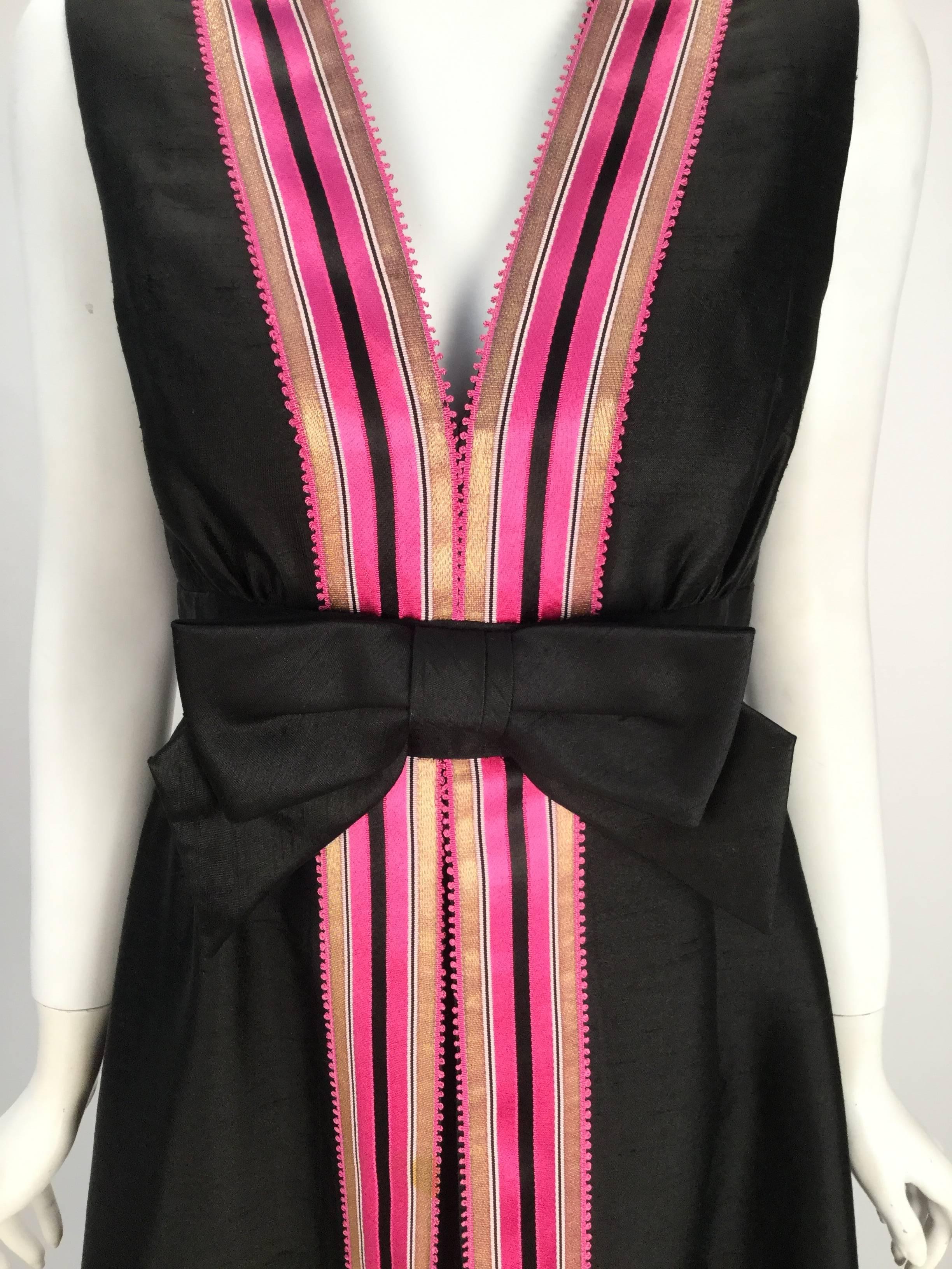 Wonderful 1960s Kent Originals Black dress featuring a fun a pink, gold and black ribbon trim descending its entire length. Front box pleat running from waist to floor. Large bow center front with a defined waist. Zip center back. Three hook and