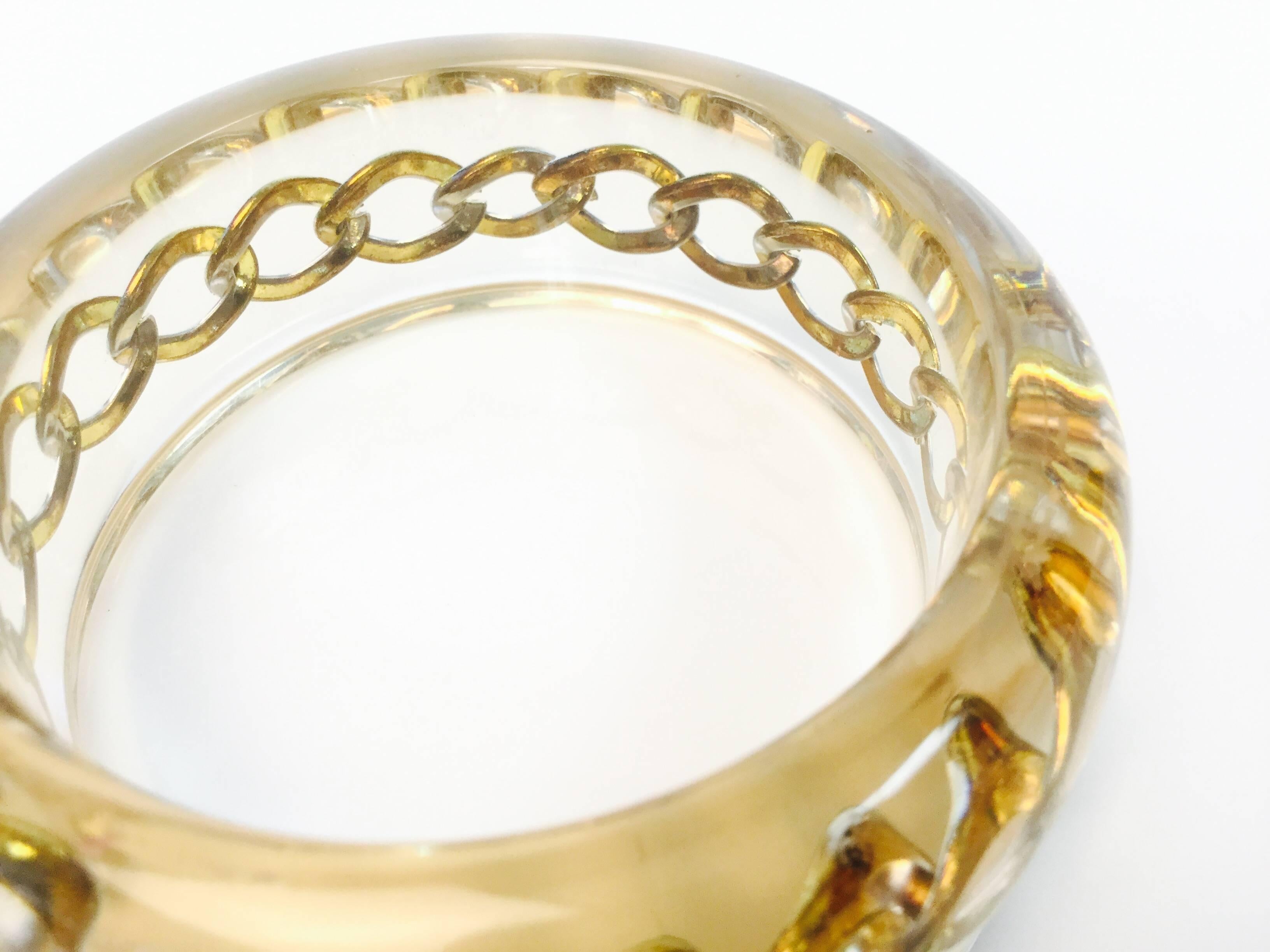 
Fantastic 1980s Lucite curb chain bangle. The bangle features a gold-tone curb chain bracelet suspended in the center of a lucite bangle. The chain links are encased in a thin bubble of air, causing the outline of the chain to lightly shimmer. The