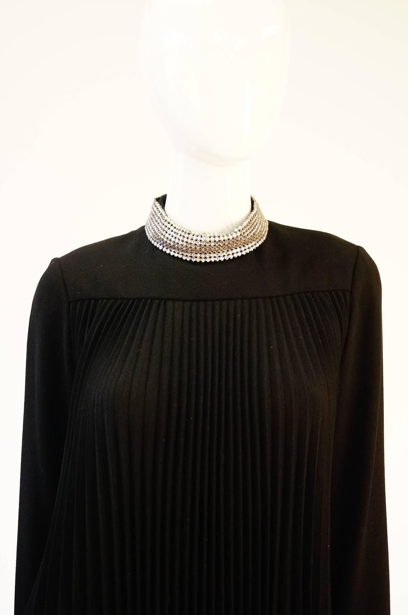 
Elegant 1960s black dress by Teal Traina New York! This graceful knee length black dress has long sleeves with shimmering rhinestone cuffs, and a rhinestone collar. The dress is primarily composed of thin pleats that sway gently with every step.