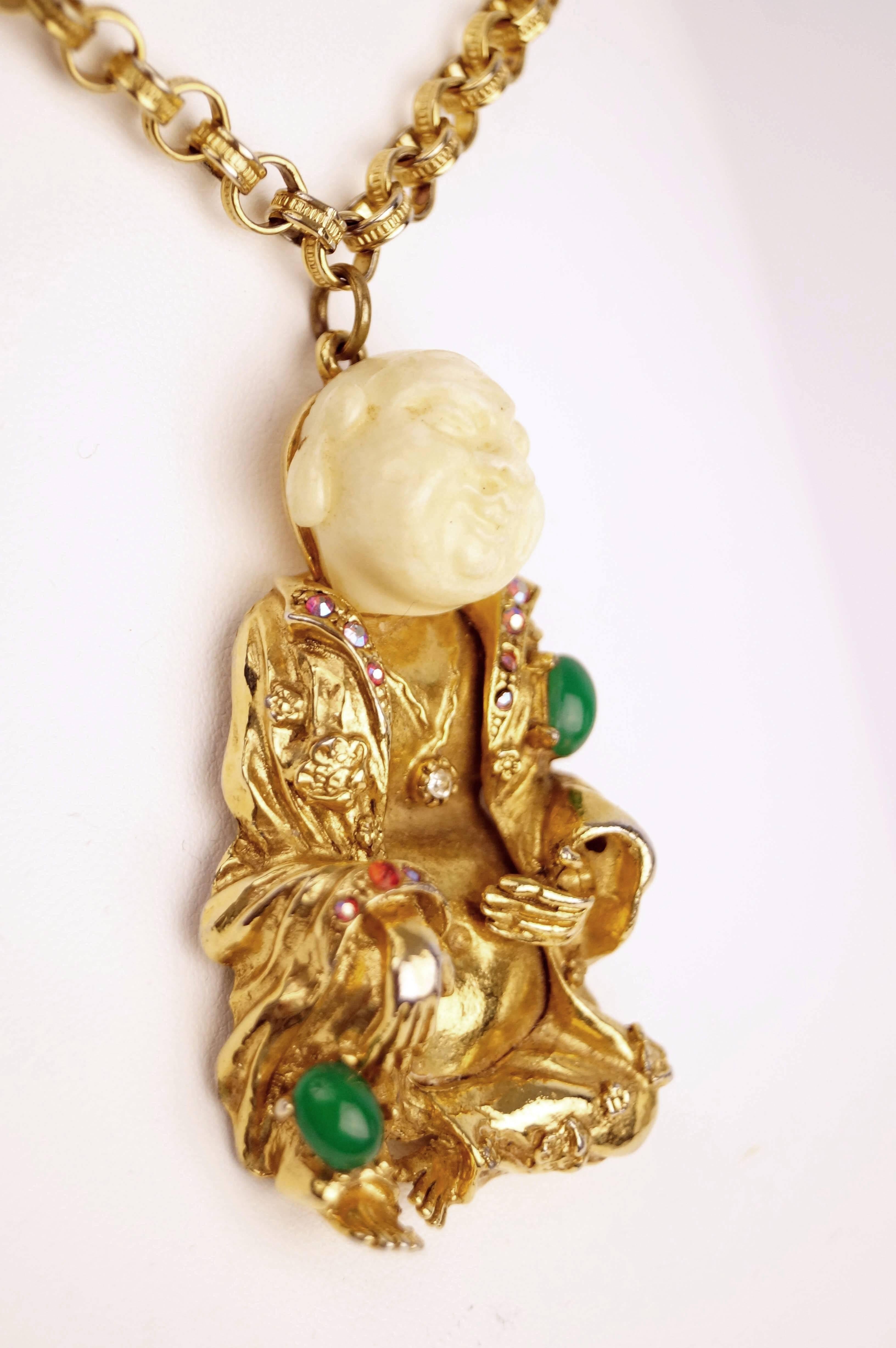 
This fun vintage laughing Buddha pendant by ART depicts a lounging figure with a faux ivory head and gold-tone body. The figure is wearing an elaborate open robe and medallion. The robe is adorned with faux jade cabochons and aurora borealis