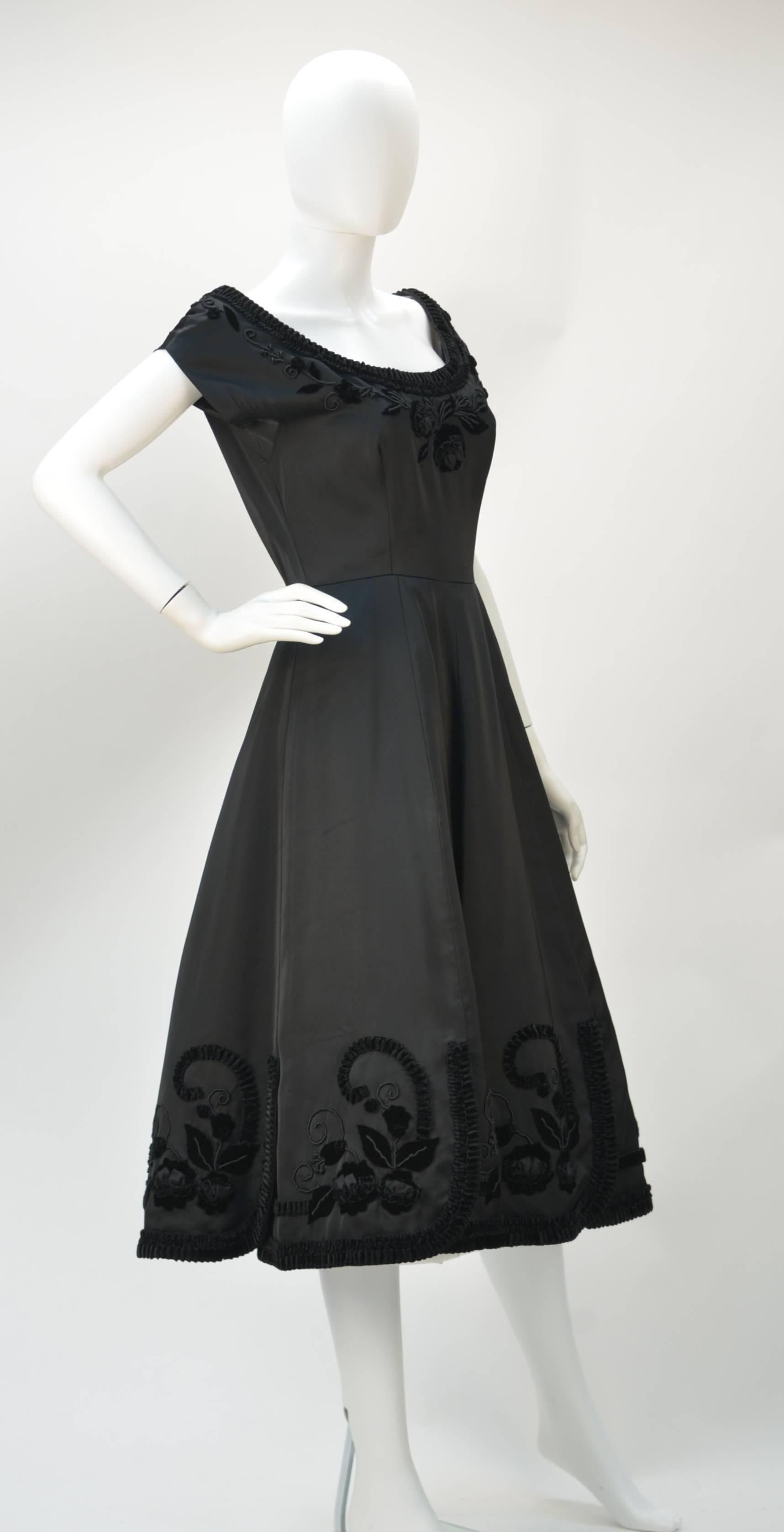 1950's black satin cocktail dress with velvet trim and excellent velvet floral detail. The dress is lined with linen and has short sleeves. The bottom of the skirt has box style pleating. The dress has a back zipper and a hook-and-eye