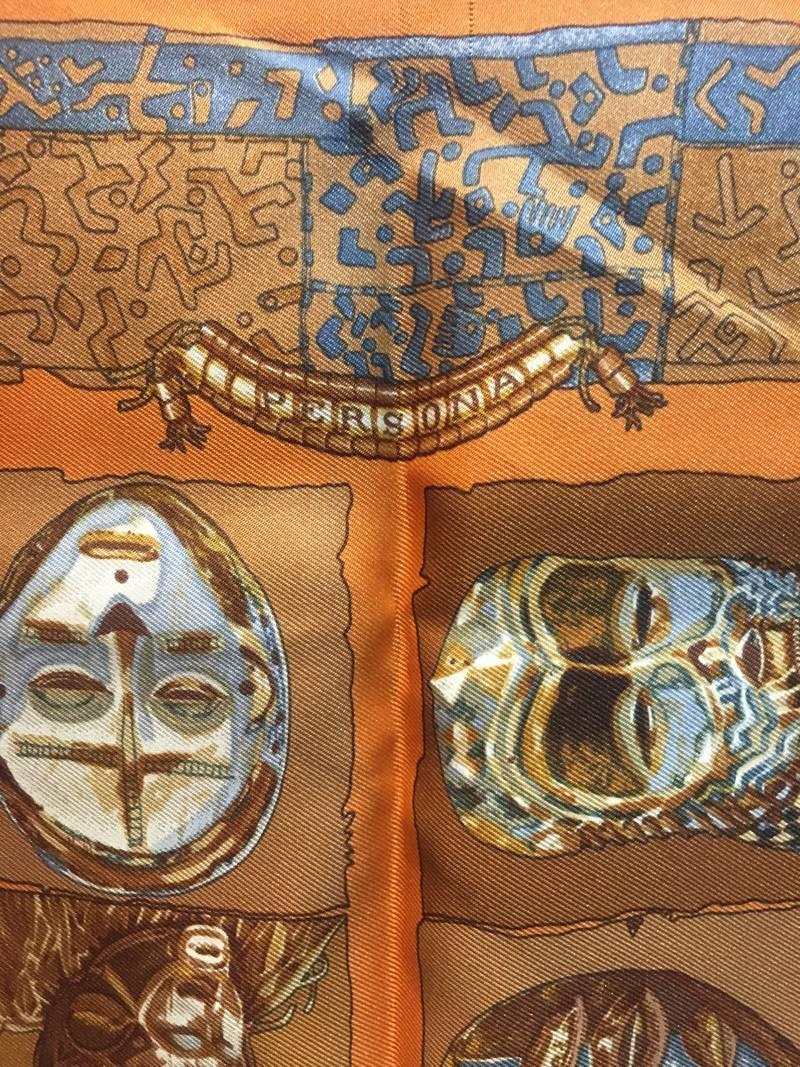 BEAUTIFUL Hermes orange personas silk pocket square scarf in excellent condition. Original silk screen design c1997 by Loïc Dubigeon features an array of traditional ancient indigenous clay masks in various neutral colors over an orange purple