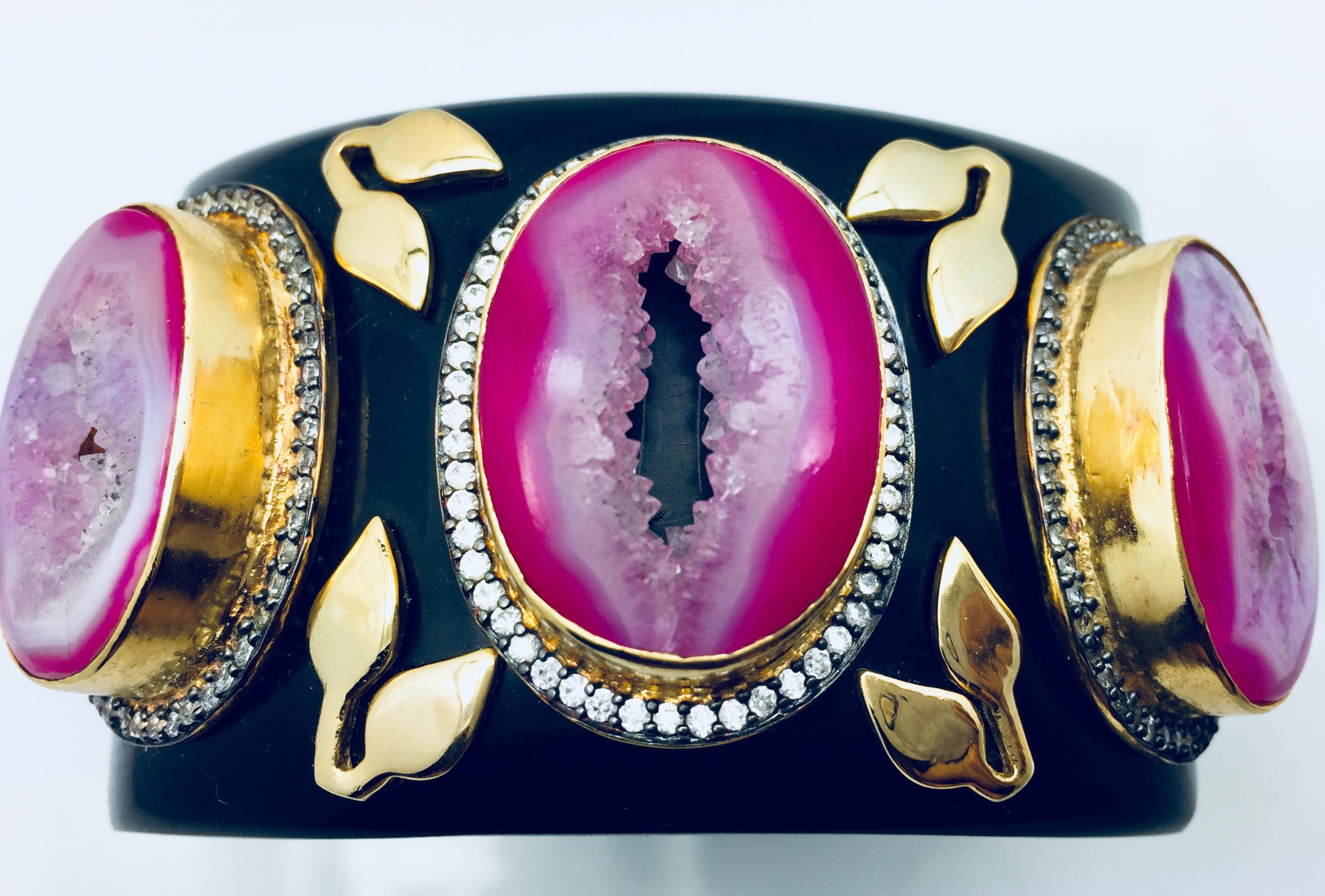 Il bracciale in resina nera liscia fa da sfondo alle pietre druzy a cratere. Ciascuno dei druzy è incorniciato da un abbagliante zircone e impreziosito da un motivo a foglie realizzato a mano.  Il bracciale è incernierato e si chiude con un inserto