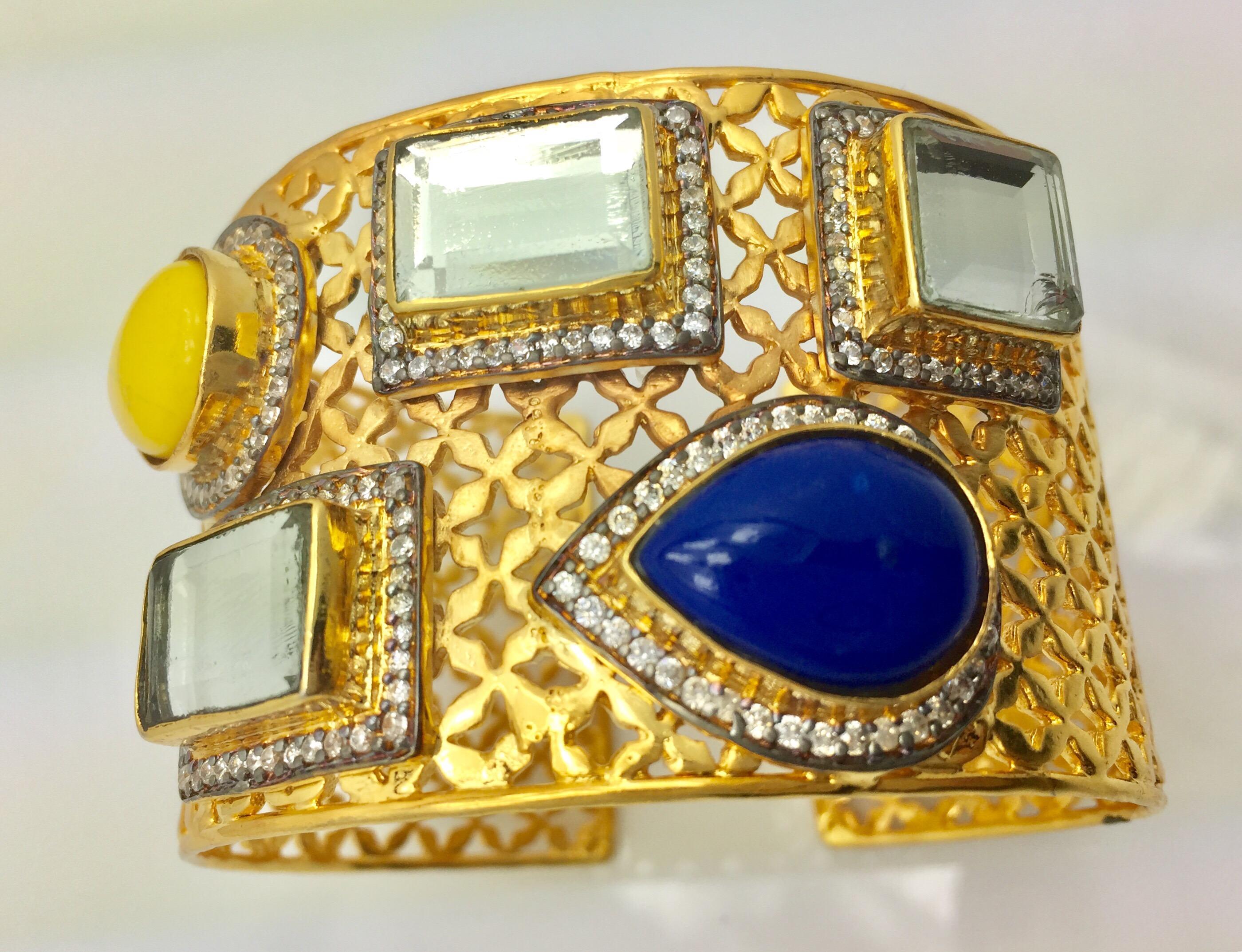 A gold frame becomes a gleaming backdrop for the brightly colored blue and yellow teardrop resin stones, enhanced with light reflecting mirror stones. The statement cuff is an attention commanding finishing touch you will treasure, and is slightly