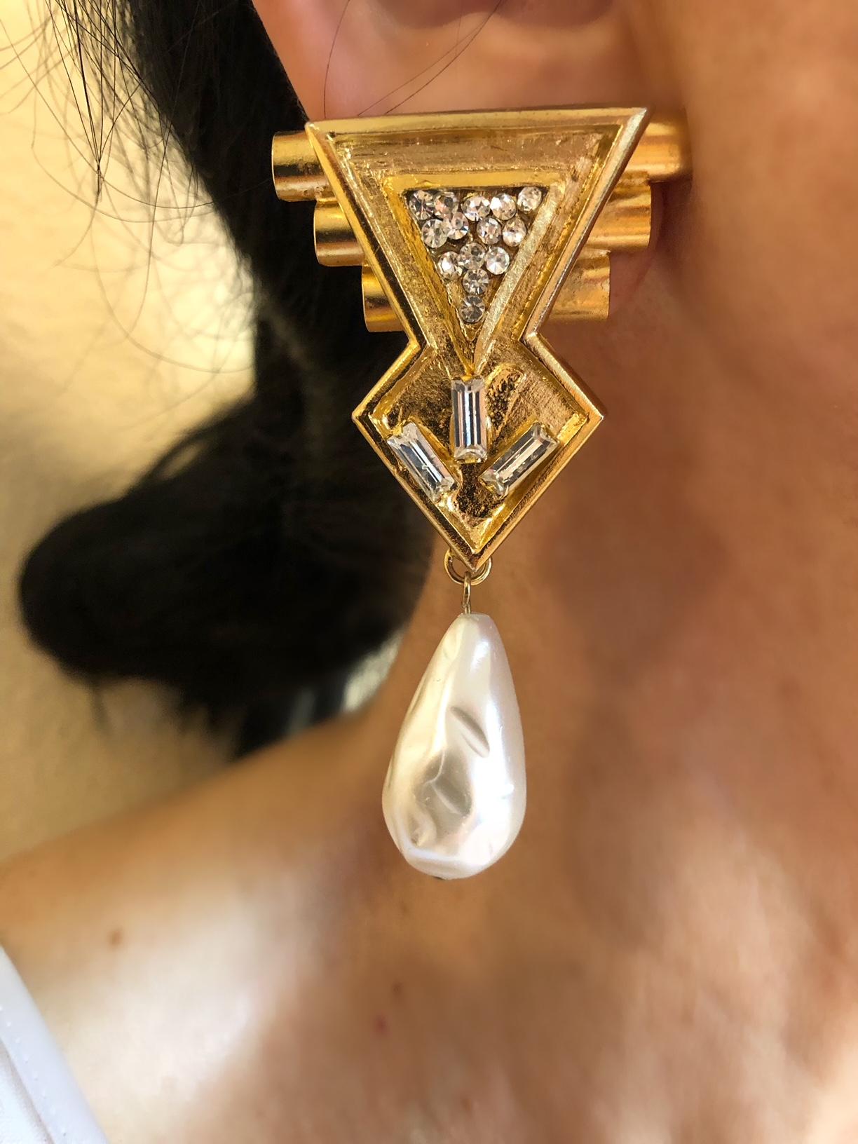 The 1980's are back, and this pair of vintage Italian clip-on earrings are no exception. Comprised of gold-tone metal featuring an edgy 1980's geometric design. The earrings are accented by clear rhinestones and large glass faux baroque dangling