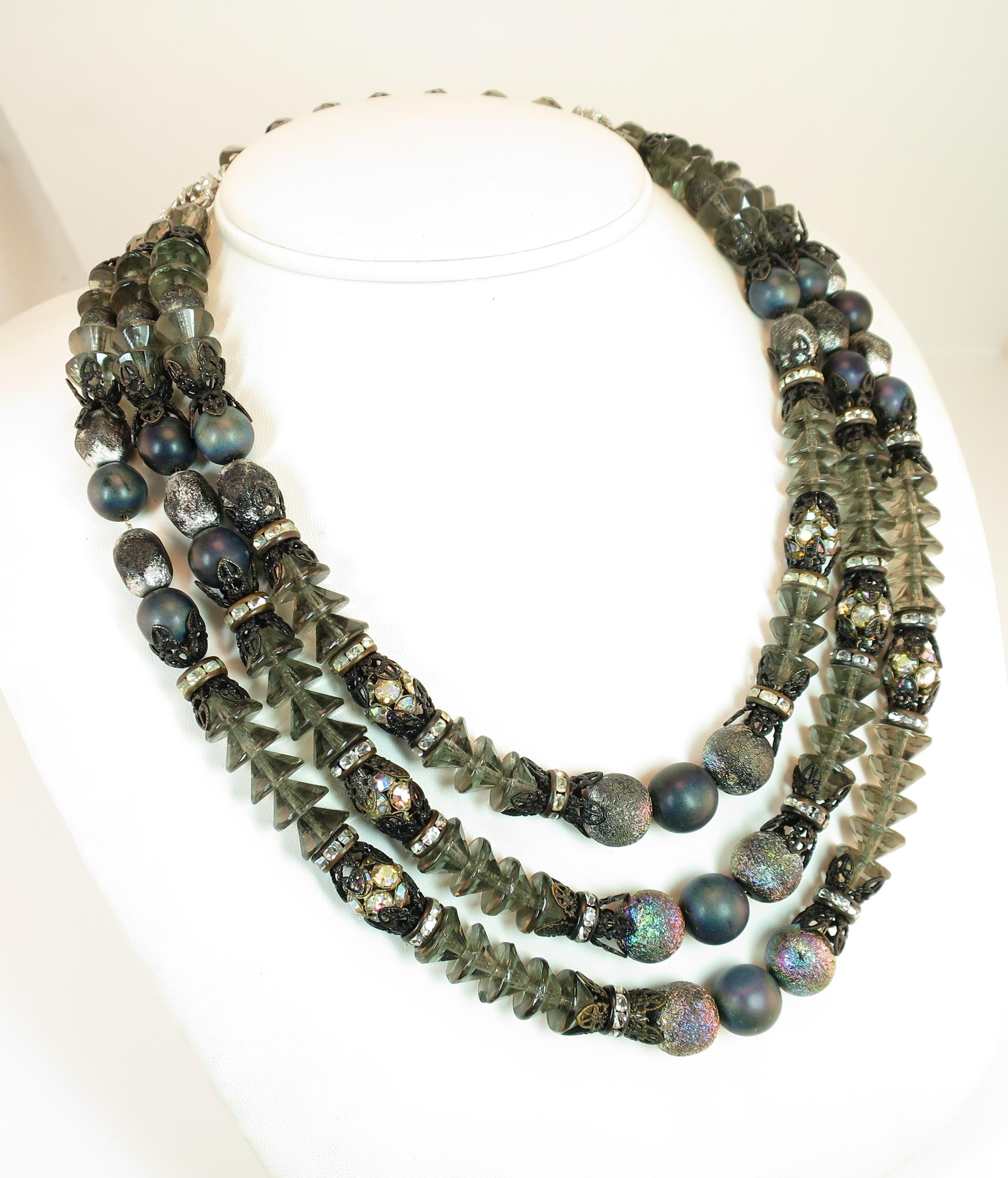 Offered here is a 1950s Hobé Mid-Century parure consisting of a three-strand draping necklace, coil memory bracelet, and clip-back earrings. All pieces are created from combinations of round composite beads in gray-purple and iridescent hues, bicone