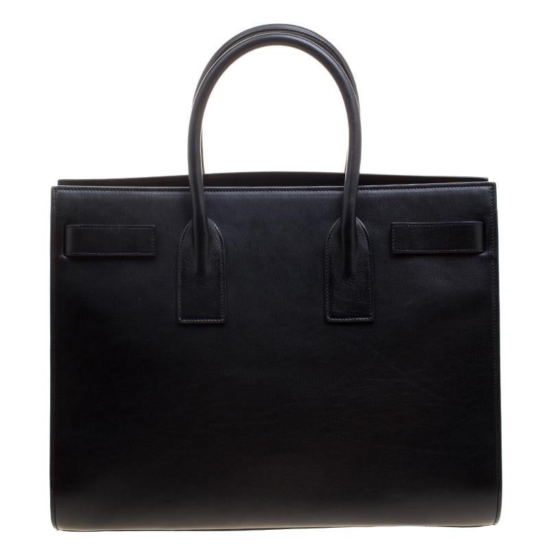 This Sac de Jour tote bag by Saint Laurent Paris has a sophisticated look. Crafted from black leather, the bag has dual round handles, a padlock and keys, and protective metal feet at the bottom. The tote comes with a suede lined interior that has