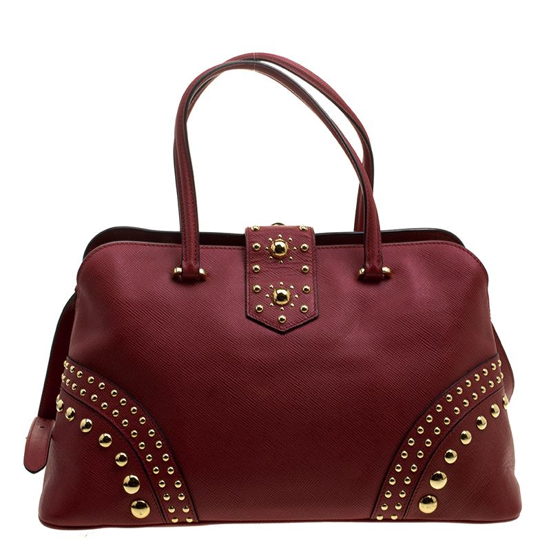 This Prada tote crafted with saffiano cuir leather in feminine red hue is perfect to complement your elegant style. It is adorned with gold-tone stud embellishments with an easy turn lock closure. The interior is leather and nylon lined featuring