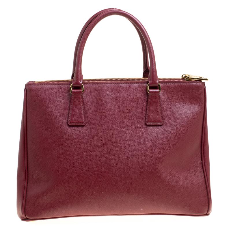 This elegant tote from Prada is crafted from Saffiano leather and is perfect for daily use. The bag features double handles, a leather covered gold key ring, detachable shoulder strap, and gold-tone hardware. It has a nylon lined interior that