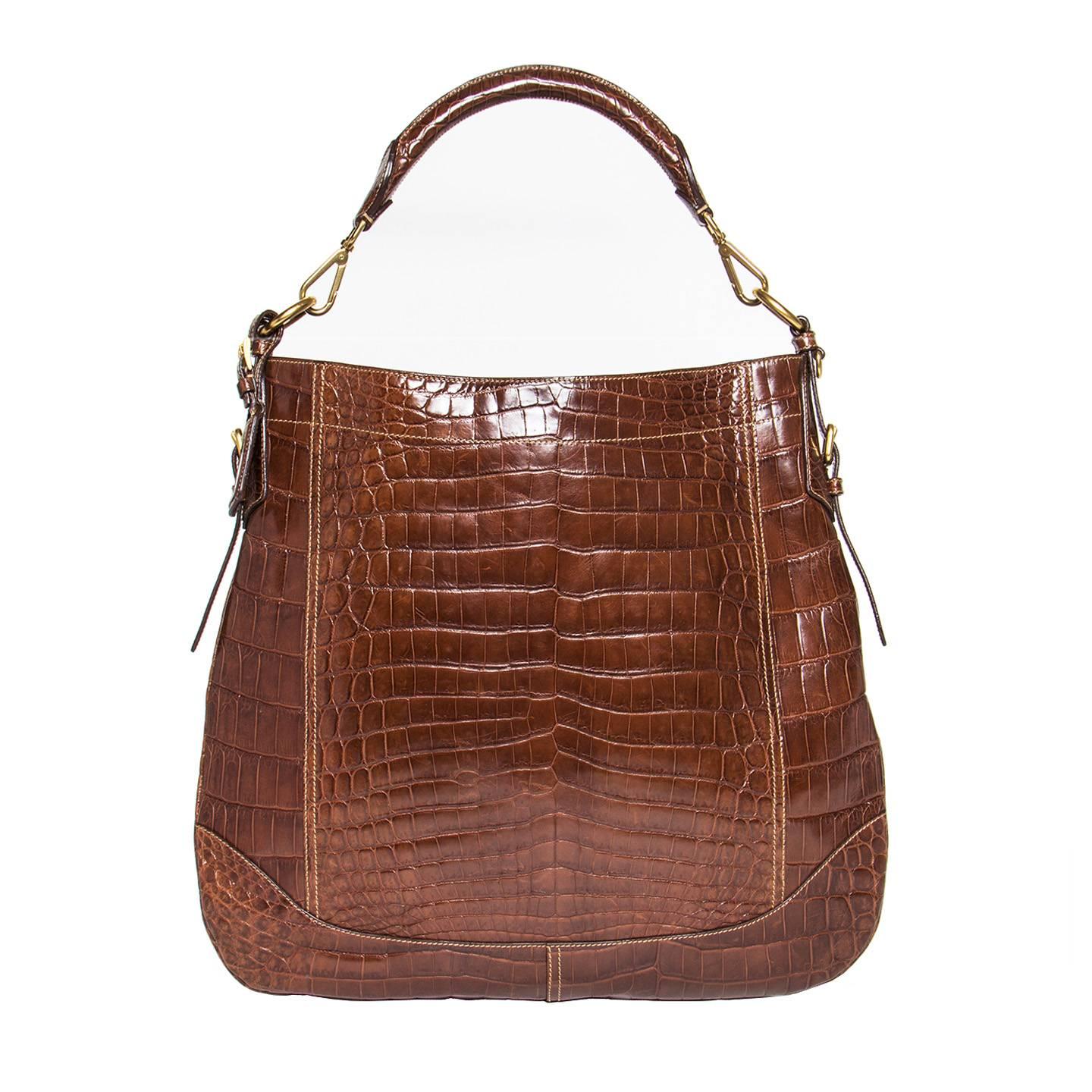 Brown crocodile medium hobo bag with Prada brushed gold metal letters at front. The sides are enriched by straps with little belt buckles and front and back are characterized by geometric panels. The shoulder strap is rounded and is attached to the