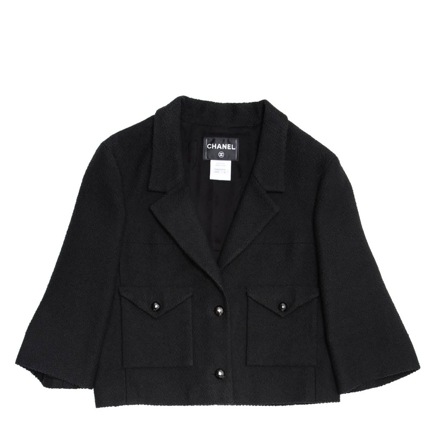 Cotton and silk boucle black cropped jacket with straight 3/4 sleeves. The jacket fastens with 2 dome shape black wrought metal buttons with a little Chanel logo, the V-neck is quite deep with a medium size lapel. Two square patch pocket with