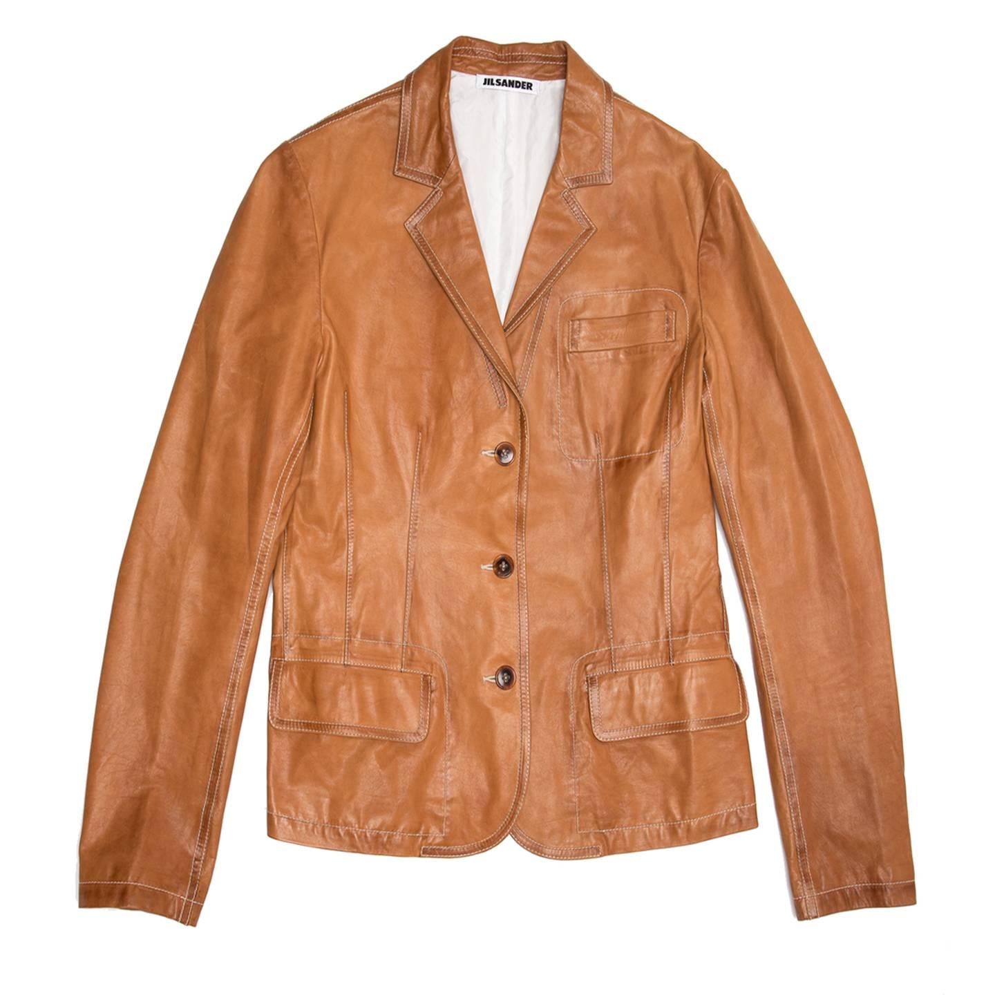 Tan brown leather short jacket with a 3 button closure and small lapel. All the seams and profiles are decorated with ivory color top stitches to match the silk lining. The front is further embellished by a breast pocket and two flap pockets, also