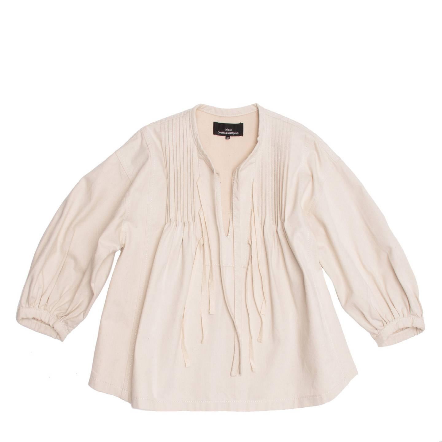 Natural color thick cotton cropped blouse with fixed pleats at top front that create an A-volume. The top is collarless and the biding on its edge ends hanging at the front. More thin ribbons dangle at the front opening. The armhole is wide and