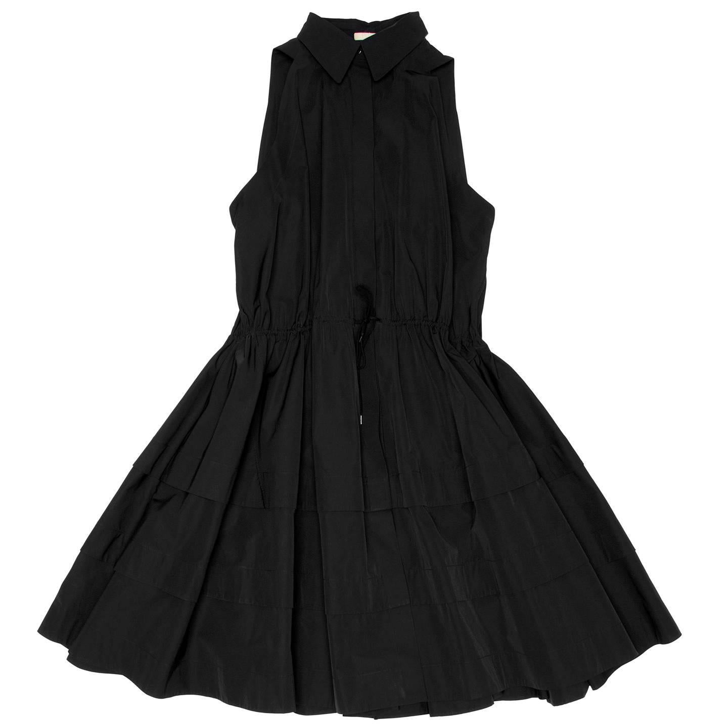 Black cotton sleeveless shirt dress with a shirt stand collar and shirred waistline. The front fastens like a shirt from top to under waist with invisible black buttons and the drawstring creates volume with gathers. The skirt is full flared and