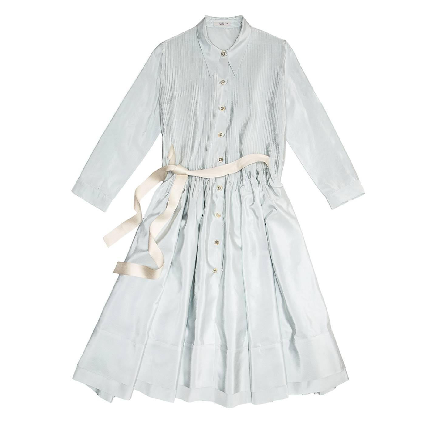 Light blue and ivory striped shiny silk shirt dress with fitted top and full skirt with below knee length. The shirt collar is long and pointed, the top part is enriched by little fixed pleats at front that open up at waistline creating volume for