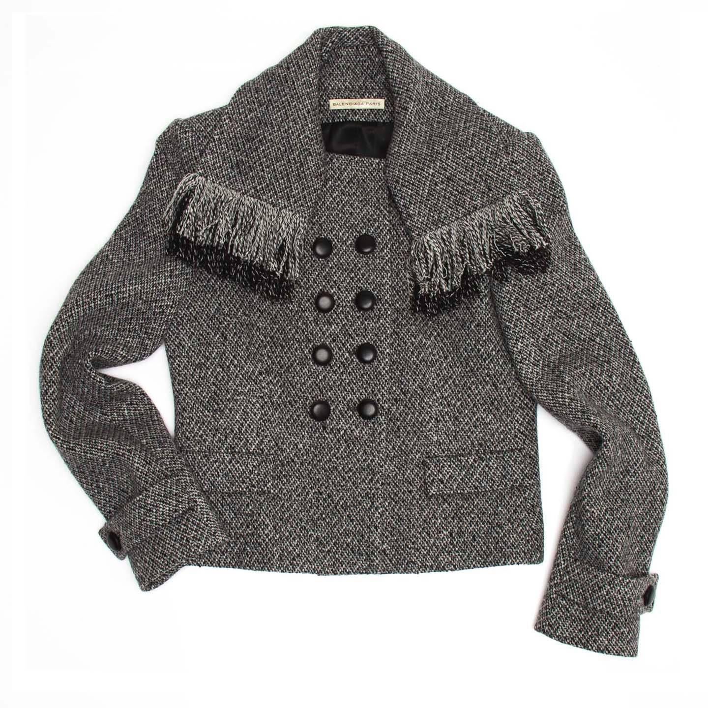 Grey tweed wool short jacket with scarf collar detail enriched by fringes. The jacket is shaped at waist and it has a small double breasted closure with black buttons. The front is enriched by flap pockets and the cuffs are belted. Made in
