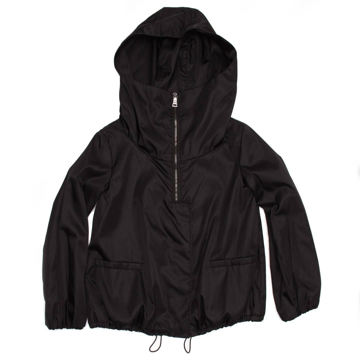 Elegant black windbreaker short jacket slightly padded, with high neck and hood. The front opens with a black metal zipper which is covered by a flap that fastens with hidden metal snap buttons. The sleeves are straight and the cuffs gathered, two