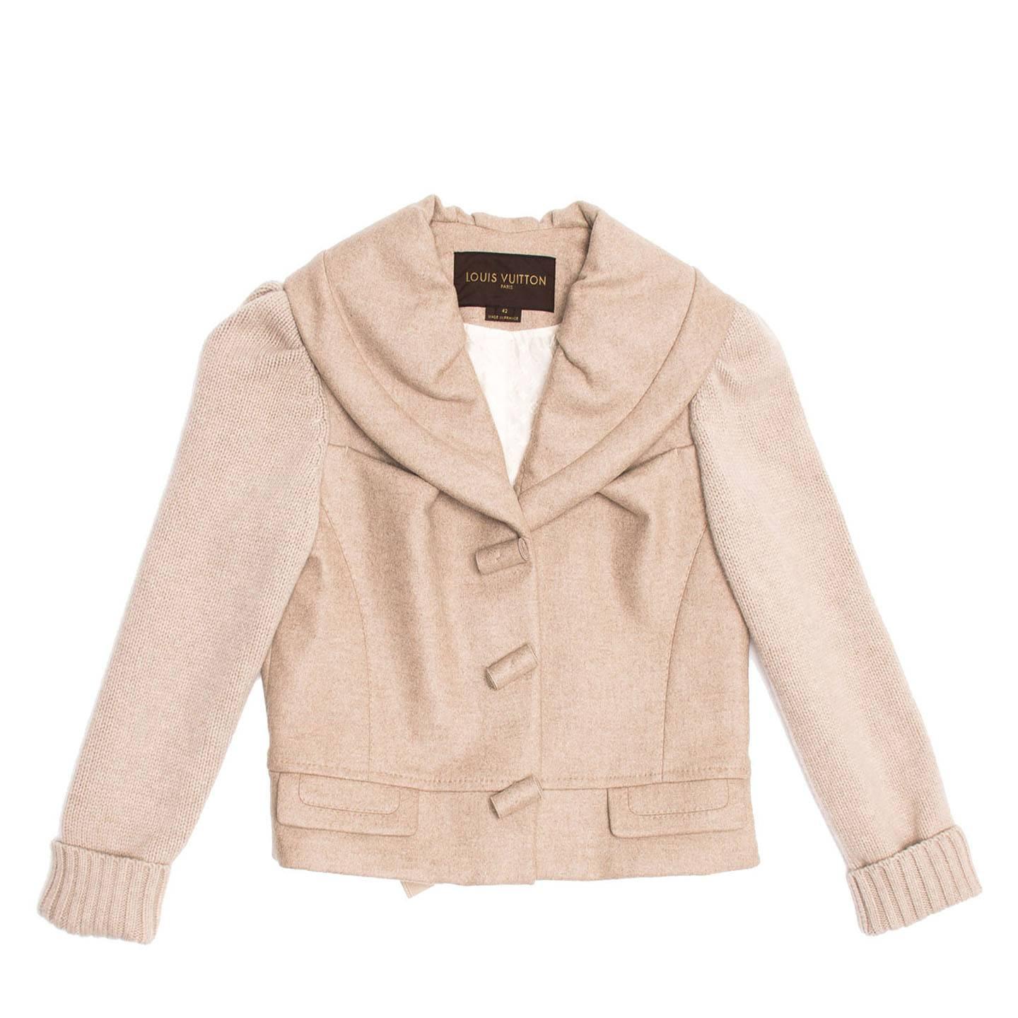Ecru waist length cashmere jacket with knit sleeves and shawl collar. Cashmere toggle style buttons fasten the front opening and two squared pockets with flaps sit at waist.The back hem width is adjustable with a ribbon tied inside the jacket and