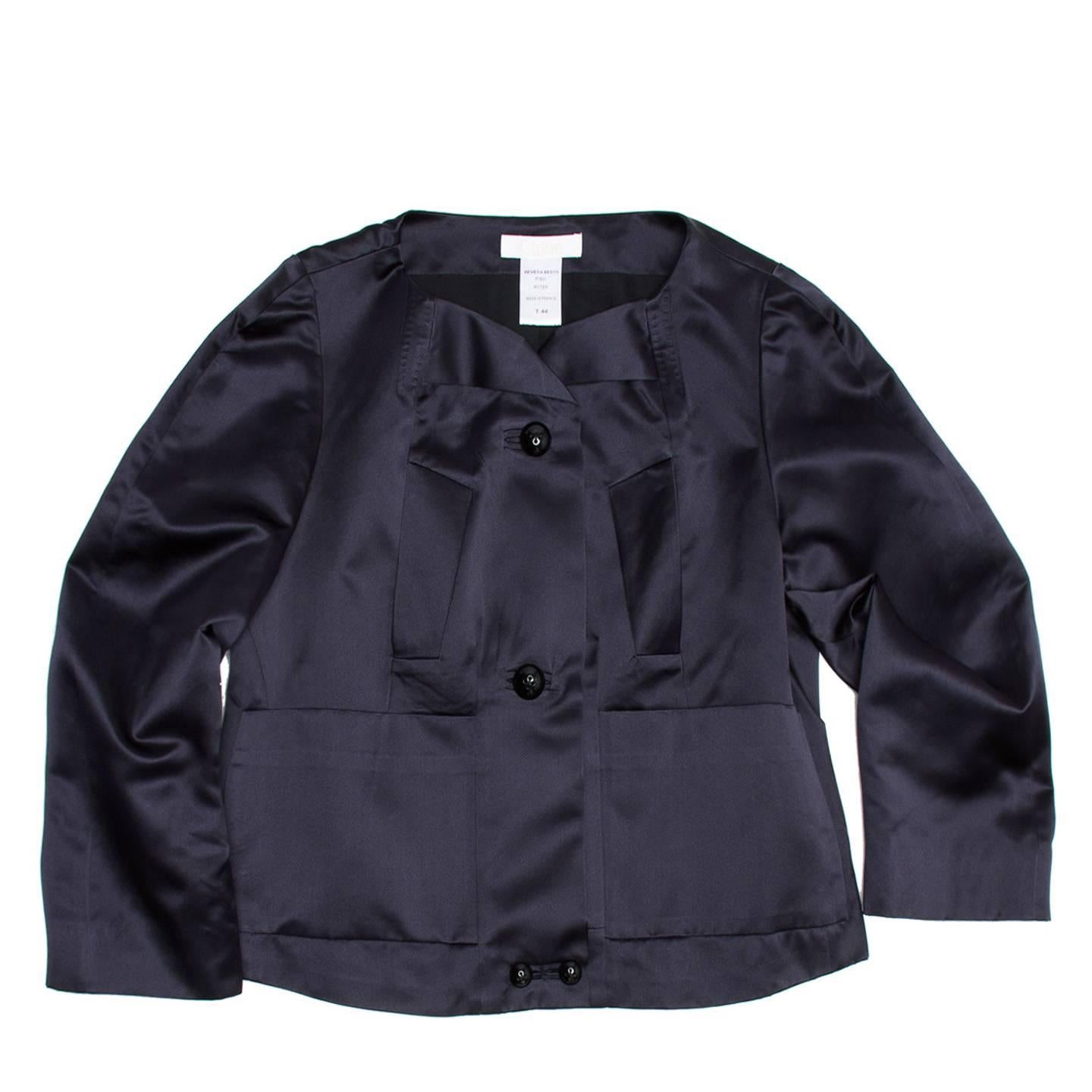 Ink silk satin collarless cropped jacket with straight fit. The front collar recalls origami folds and is enriched on the sides by tone-on-tone hand top stitches. Two slash pockets panels and two rectangular patch pockets contribute to the geometric