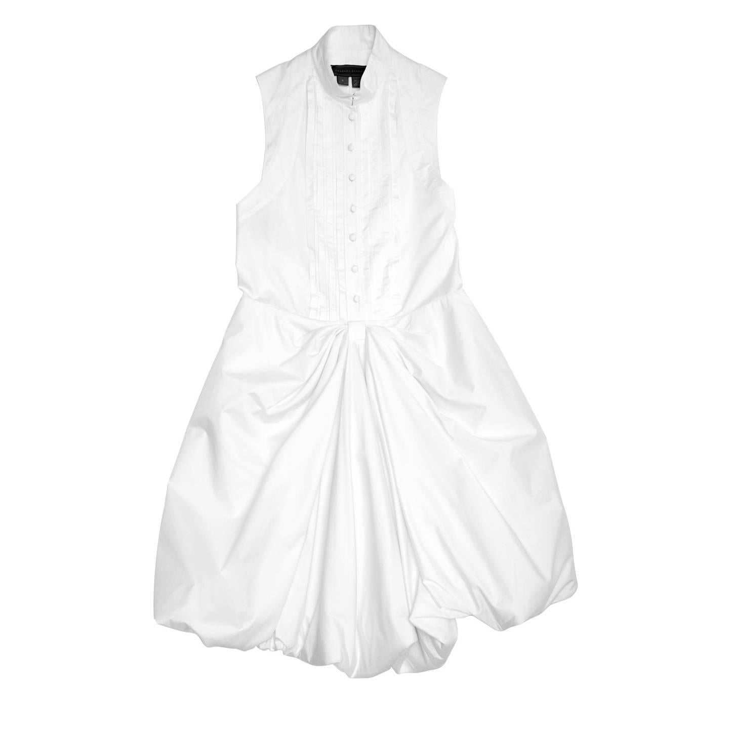 White cotton sleeveless shirt dress with full knee length puff skirt and bow style drapes at front under waist. The front fastens with covered buttons until the waist line, the bib detail is enriched by fixed pleats and the Nehru collar fastens with