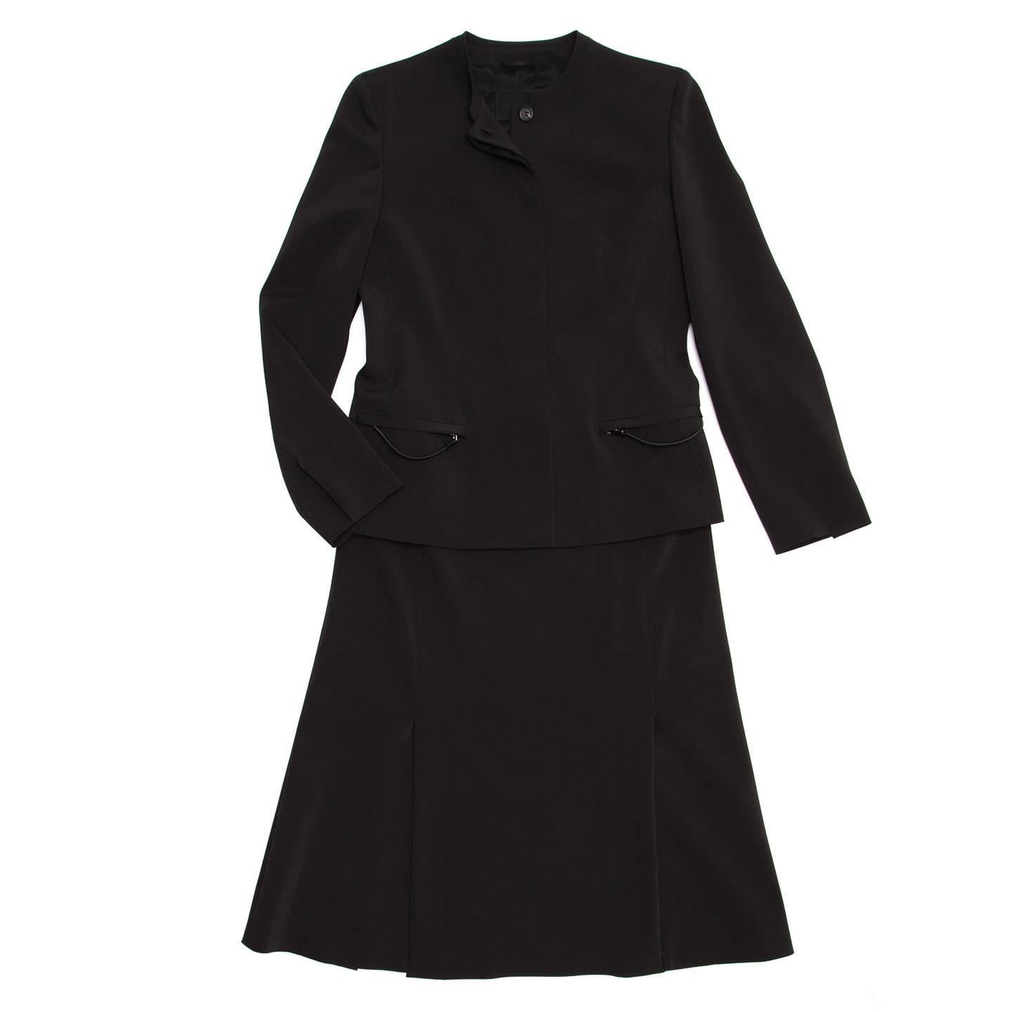 Black acetate blend skirt suit with zigzag stitching details. The jacket is hip length, has a classic round neck, a front opening with a flap covering the buttons and pockets with zippers and puller detail. The skirt is knee length, fitted until