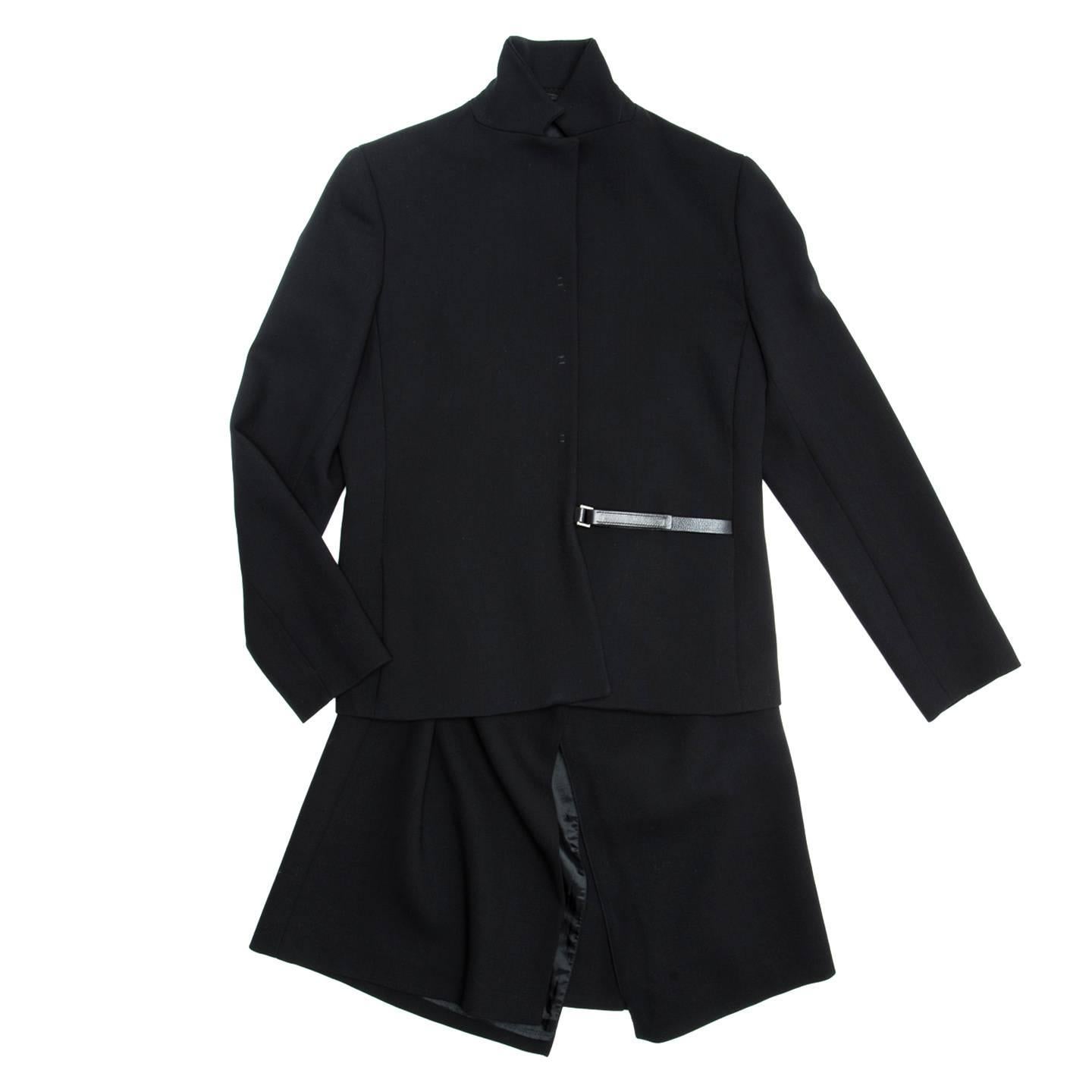 Black virgin wool skirt suit with leather detail. The jacket is hip length, has a small notched collar, vertical slit pockets, a 3 covered snap buttons front opening with tack details and a leather strap and buckle closure at waist line. The skirt