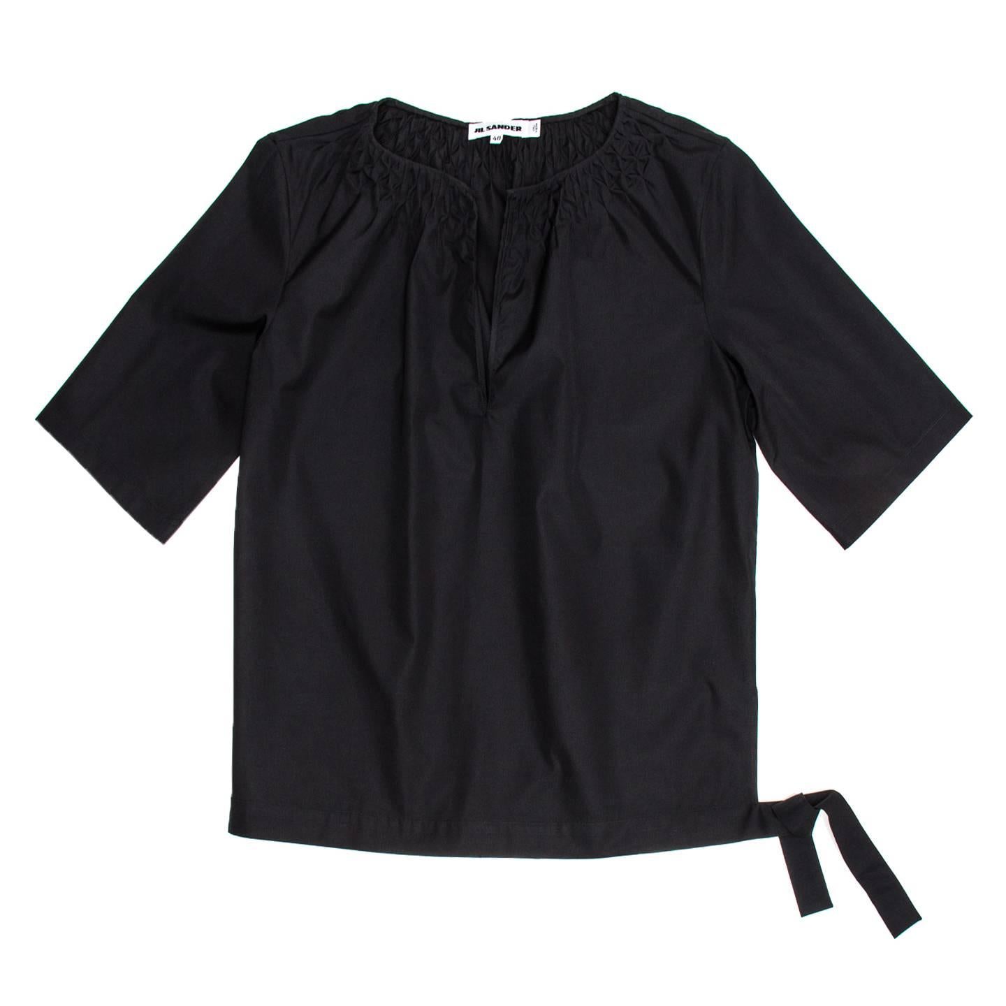 Black cotton tunic top with round collar and straight sleeves short to the elbow. The neckline opens at center front and is embellished by invisible tacks that create a geometric design at front and back, while a waistband fastens on a side with a