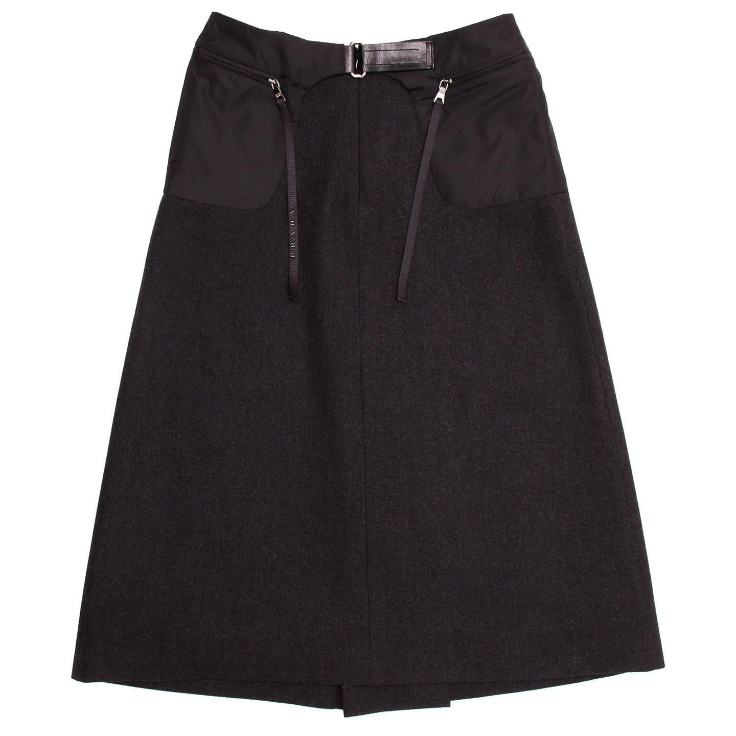 Prada Charcoal grey wool A-line skirt with black nylon round patch pockets on top-sides that open at front with metallic zippers adorned by dangling ribbons. The pockets join at front waist with a black leather insert that fastens with a silver